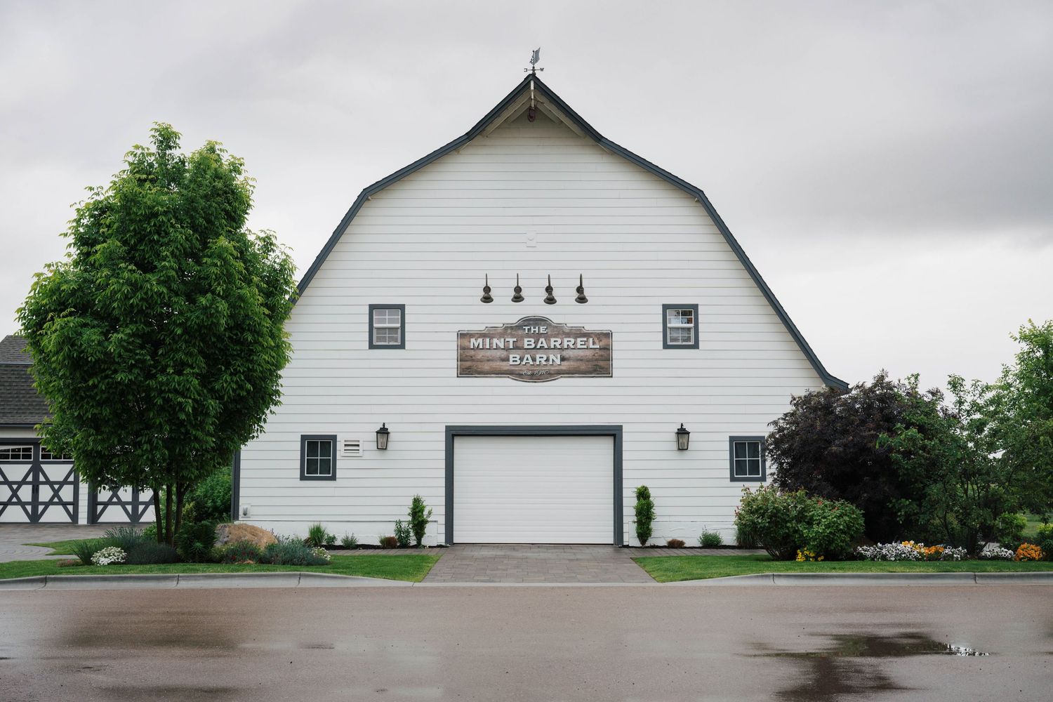 A panoramic view of a white barn with green landscaping and trees under an overcast sky.
