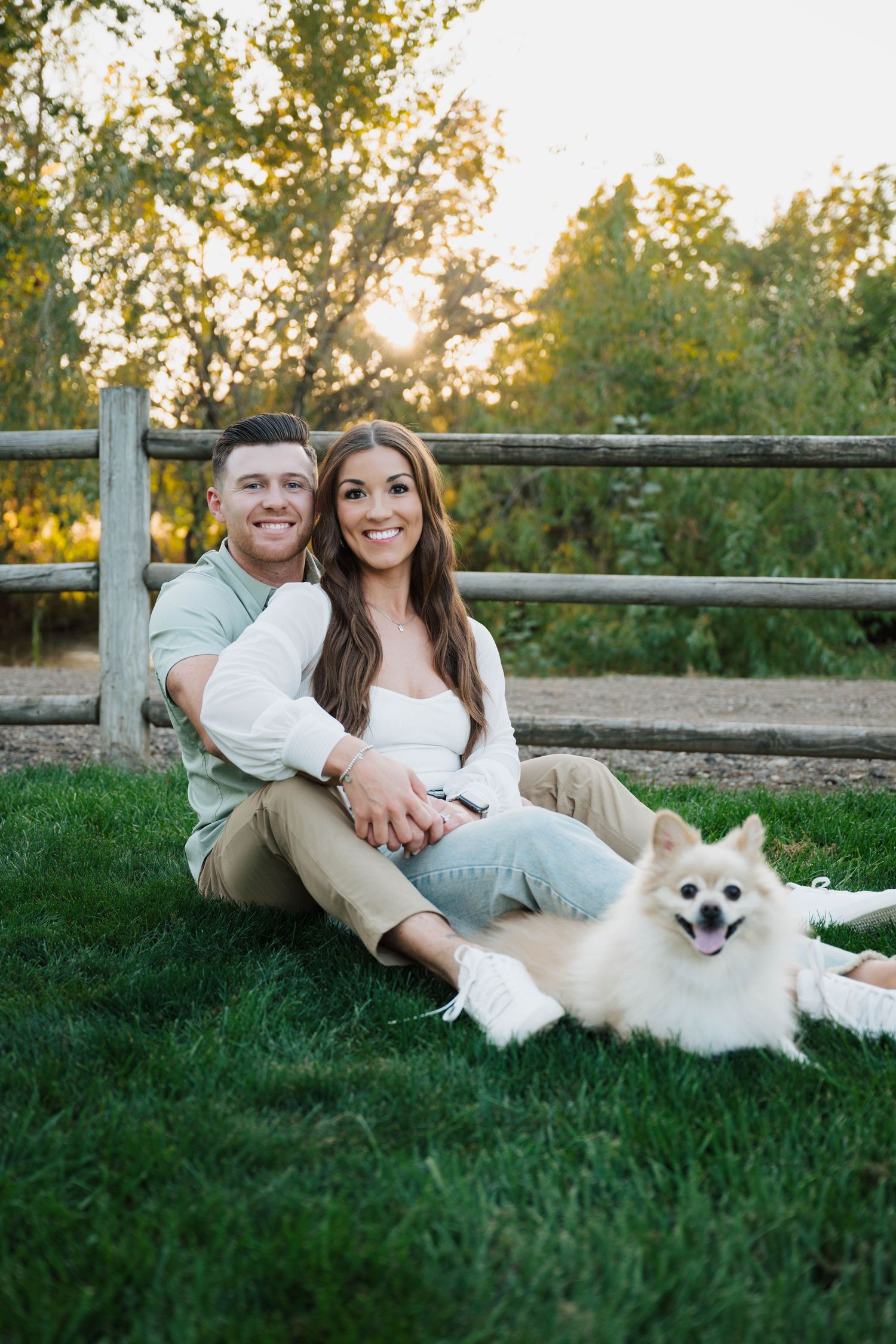 A couple relaxes on grass with their white Pomeranian dog near a wooden fence.