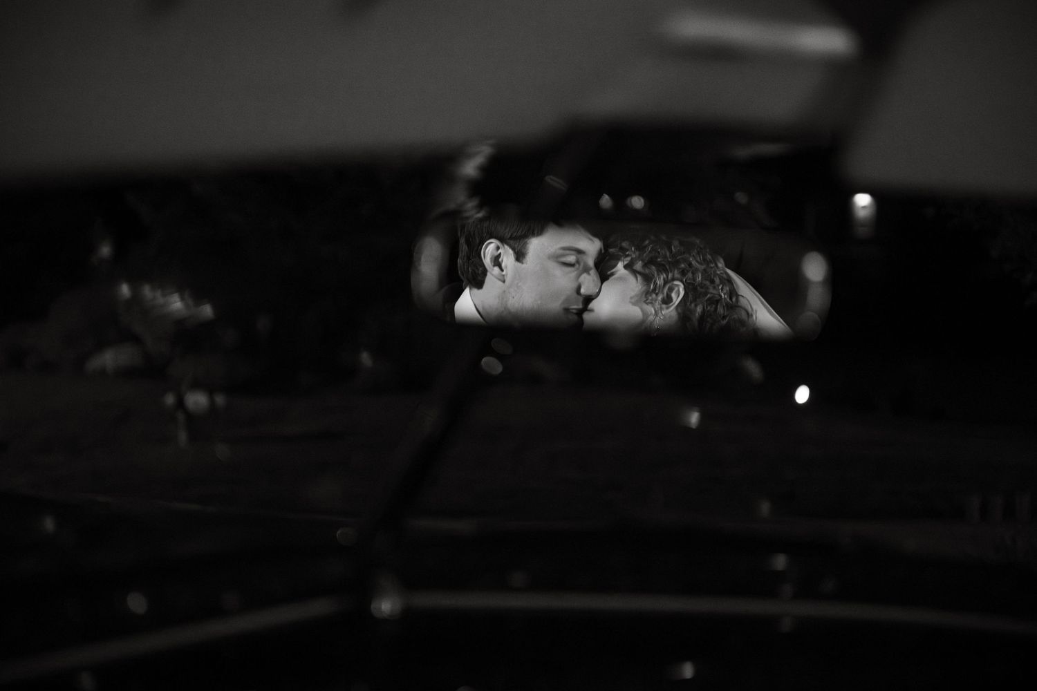 Black and white photo of a couple kissing, reflected in a car mirror at night.