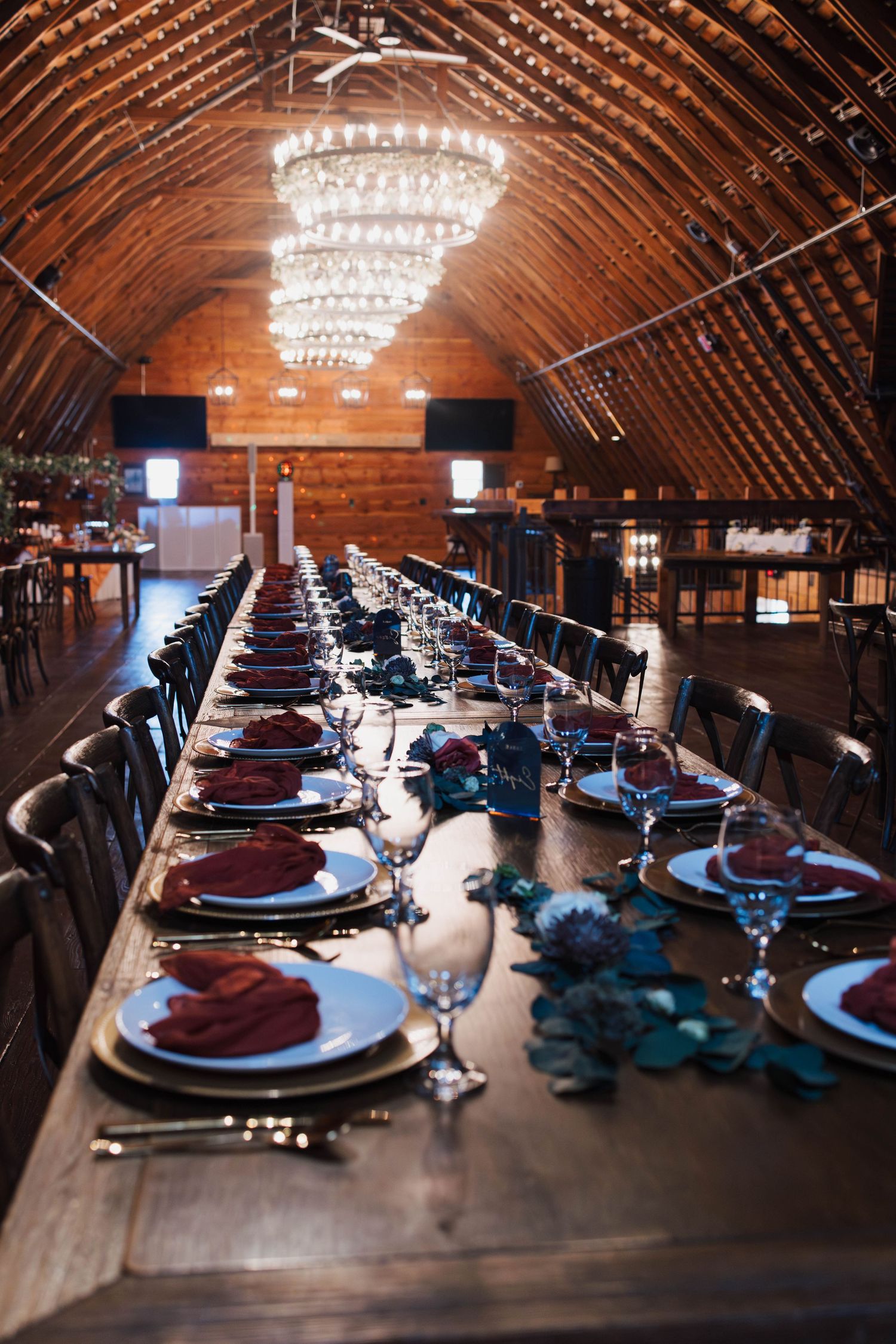 Elegant barn wedding reception with long wooden table, crystal chandelier, burgundy napkins, and blue floral decor under vaulted ceiling.