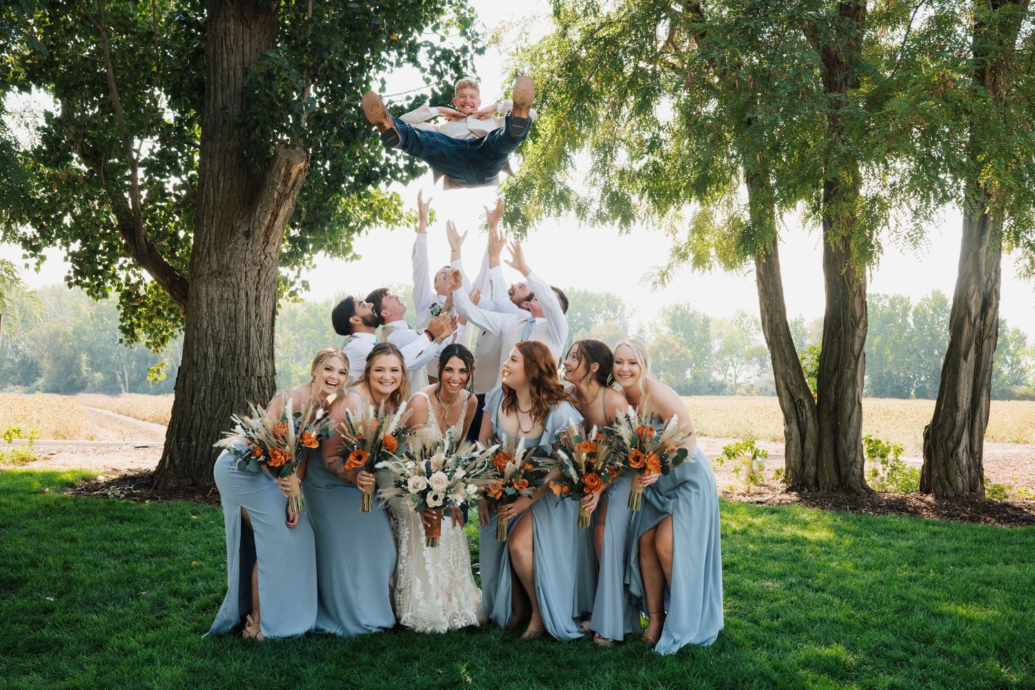 Wedding party poses outdoors, bridesmaids in sage gowns holding bouquets while groomsmen toss groom in the air.