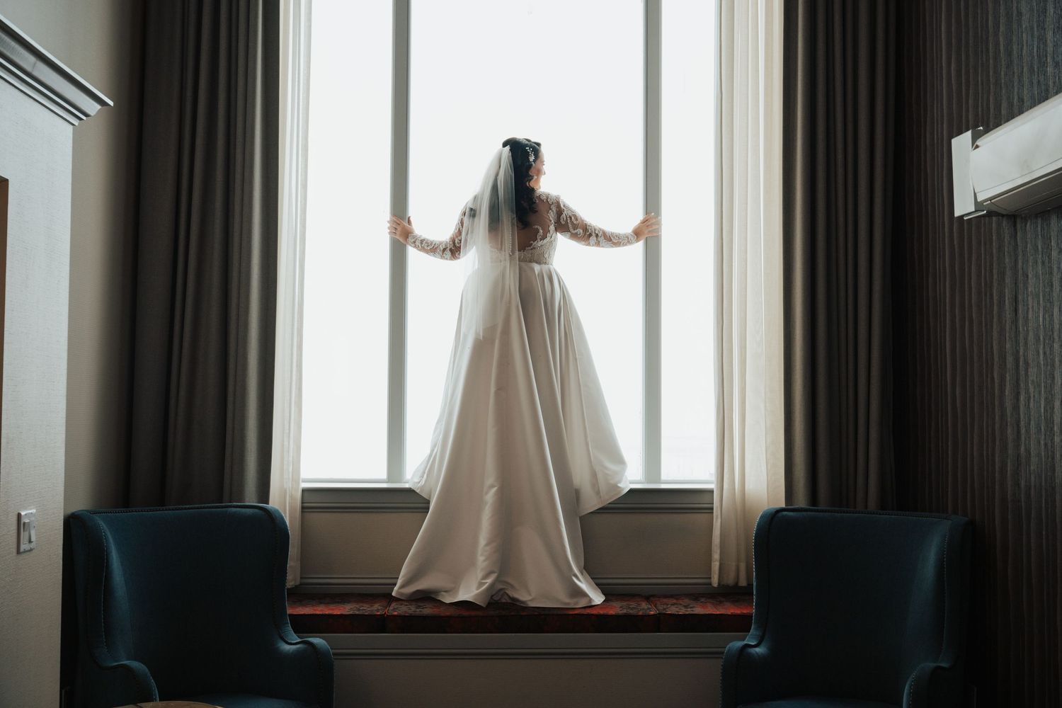 A flowing white wedding dress silhouetted against large windows with dark curtains.