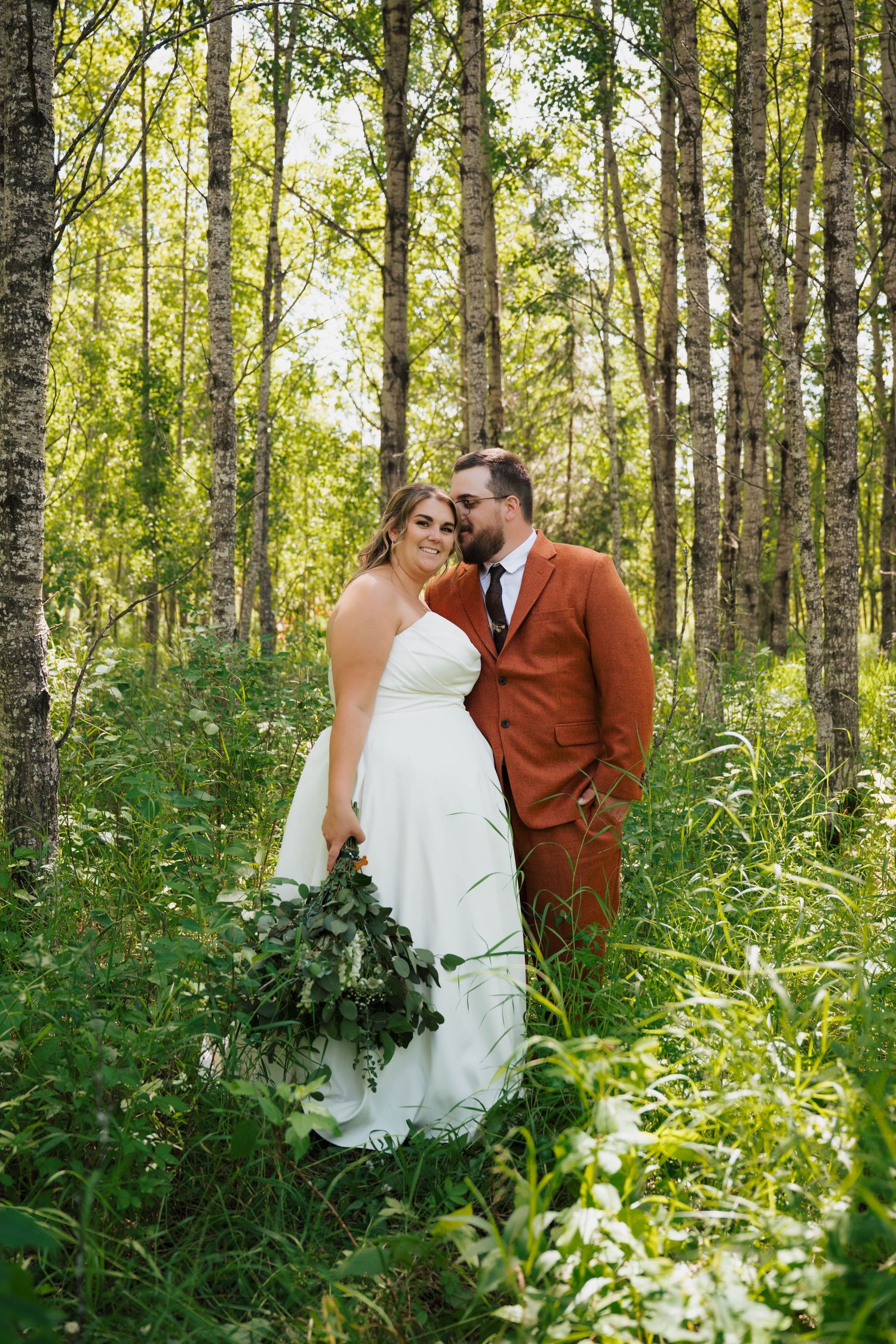 A wedding couple poses in a sun-dappled birch forest with lush greenery.