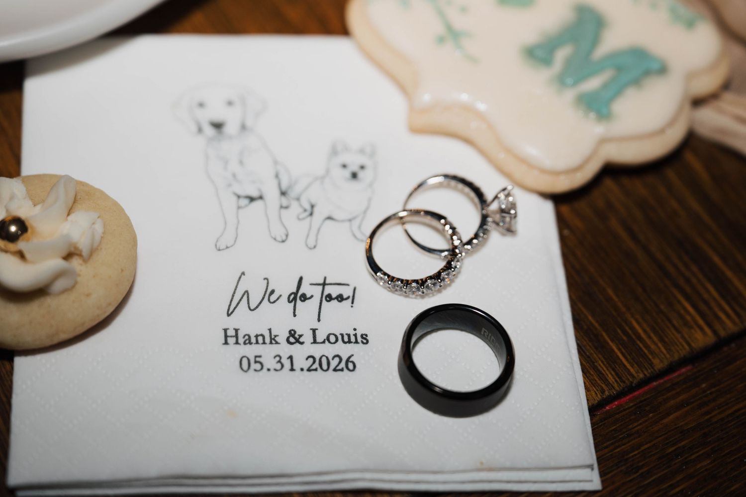 Wedding rings and bands displayed on a napkin reading We Do Too Hank and Louis 05.31.2026 with dog illustrations.