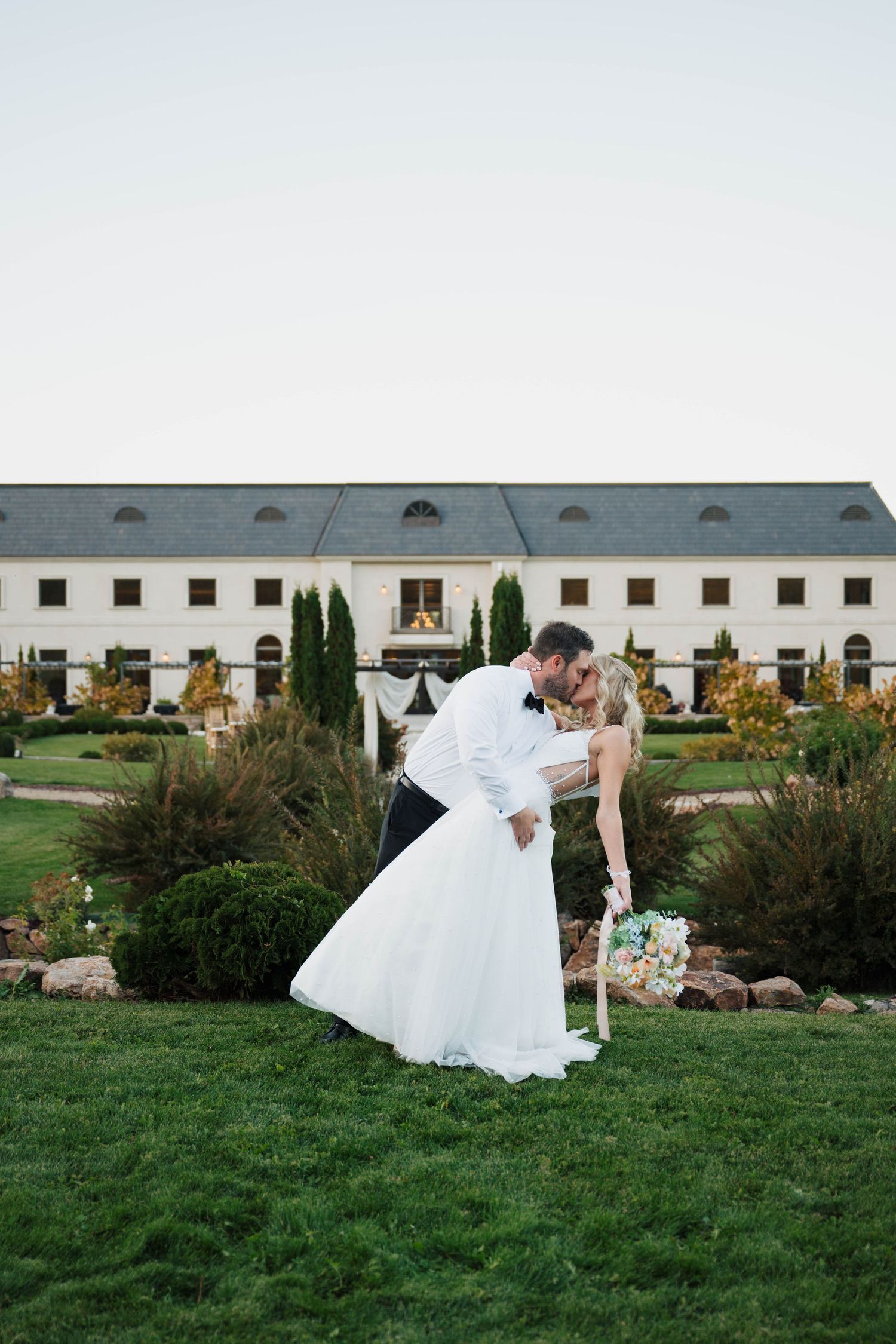 Bride and groom share a romantic dip kiss on green lawn before elegant white wedding venue at dusk.