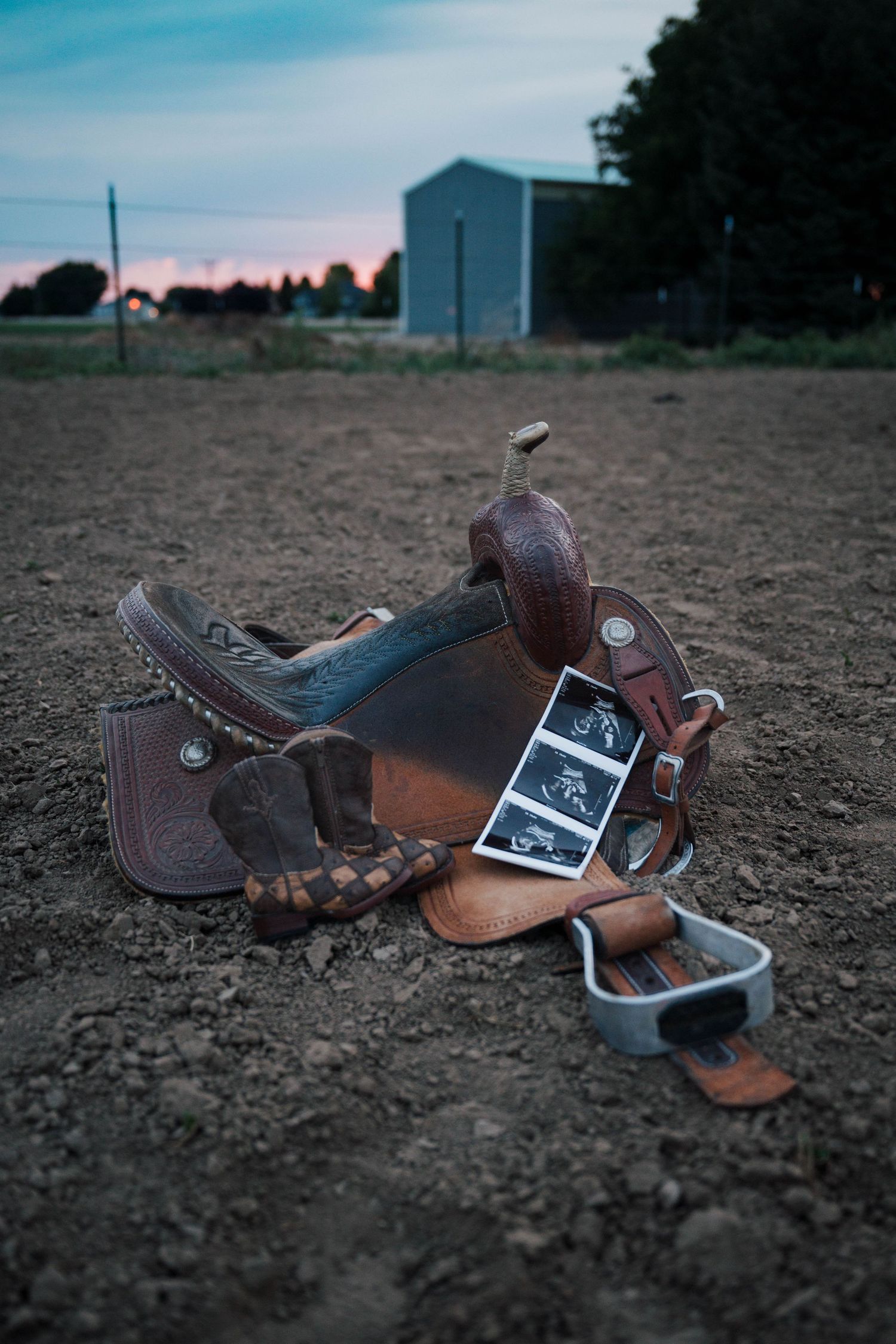 Western saddle with baby ultrasound photos tucked inside, resting on dirt ground at dusk, creative cowboy pregnancy announcement.