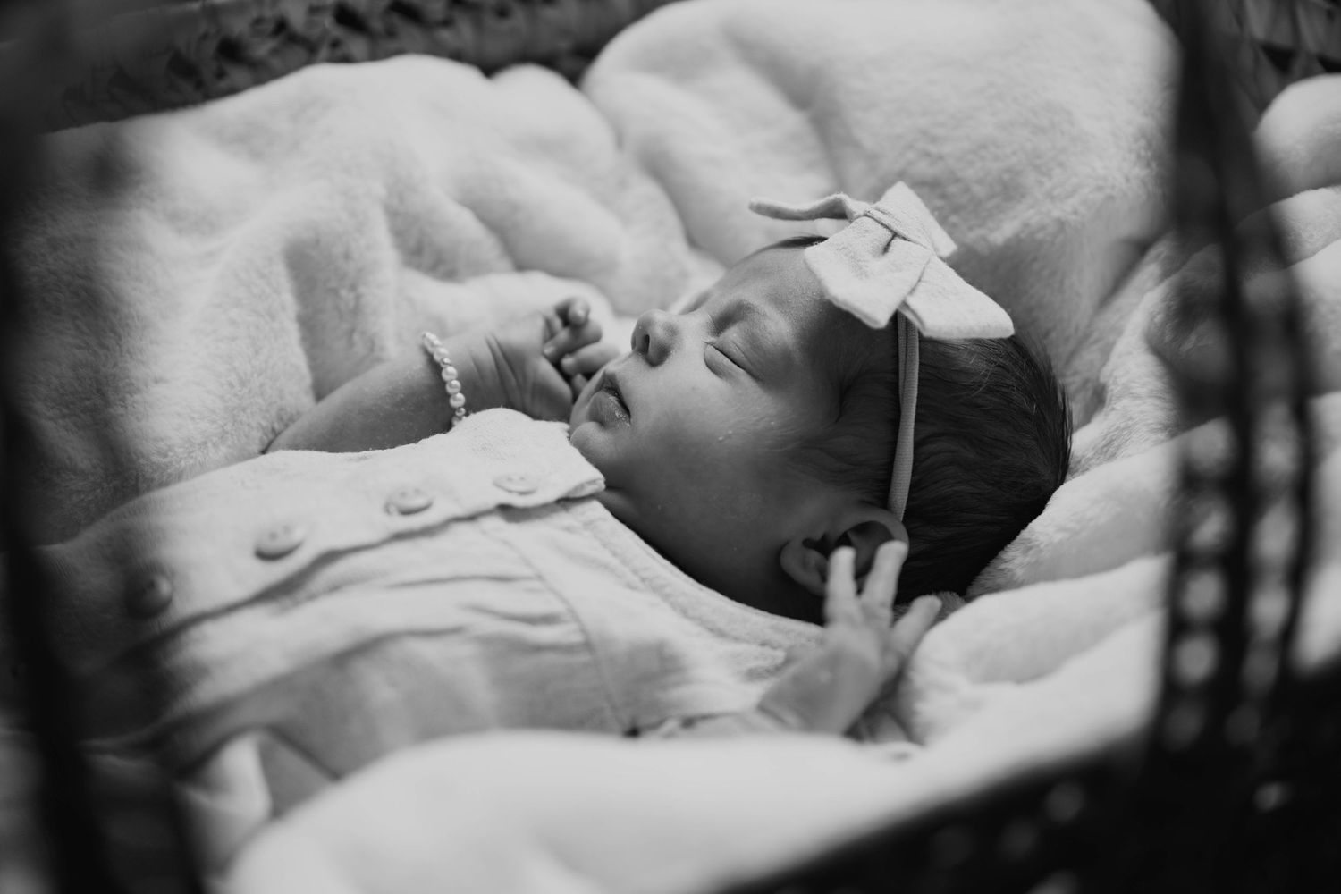 Black and white photo of a sleeping newborn baby girl wearing a bow headband in a basket.