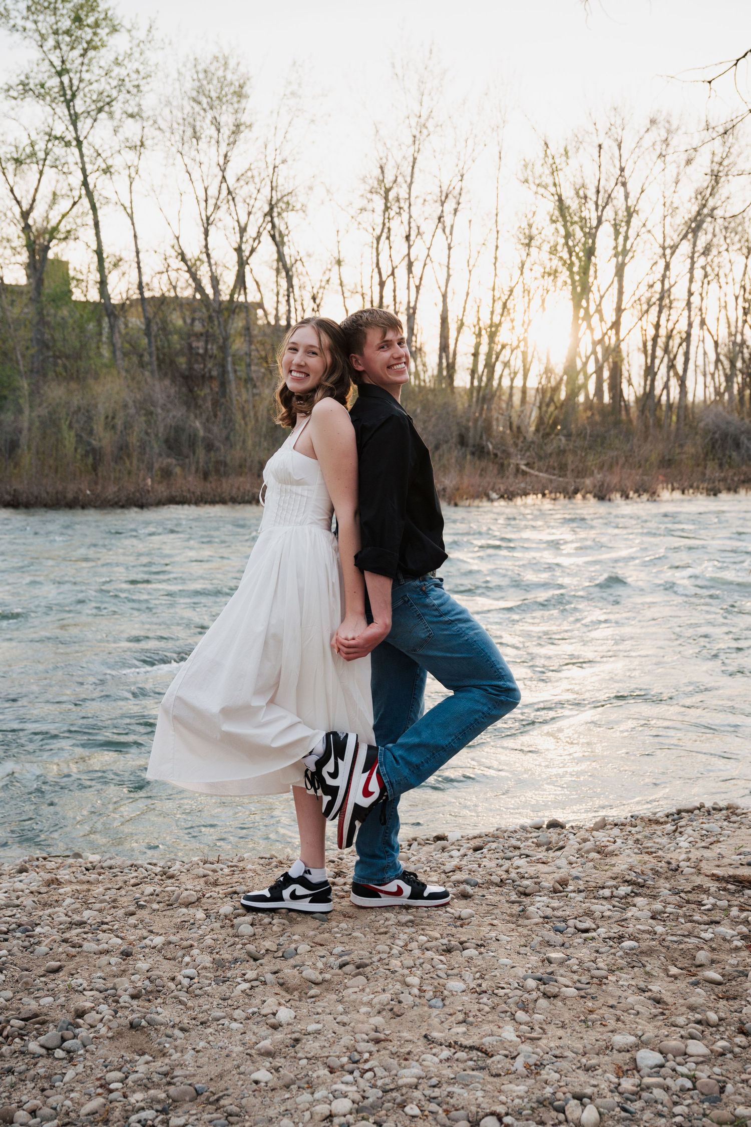 Couple in casual wear posing together on rocky riverbank at sunset.