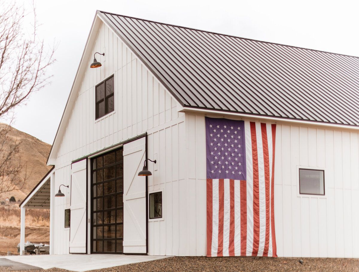 A modern white barn with a large American flag hanging on its exterior and metal roof against a desert landscape.