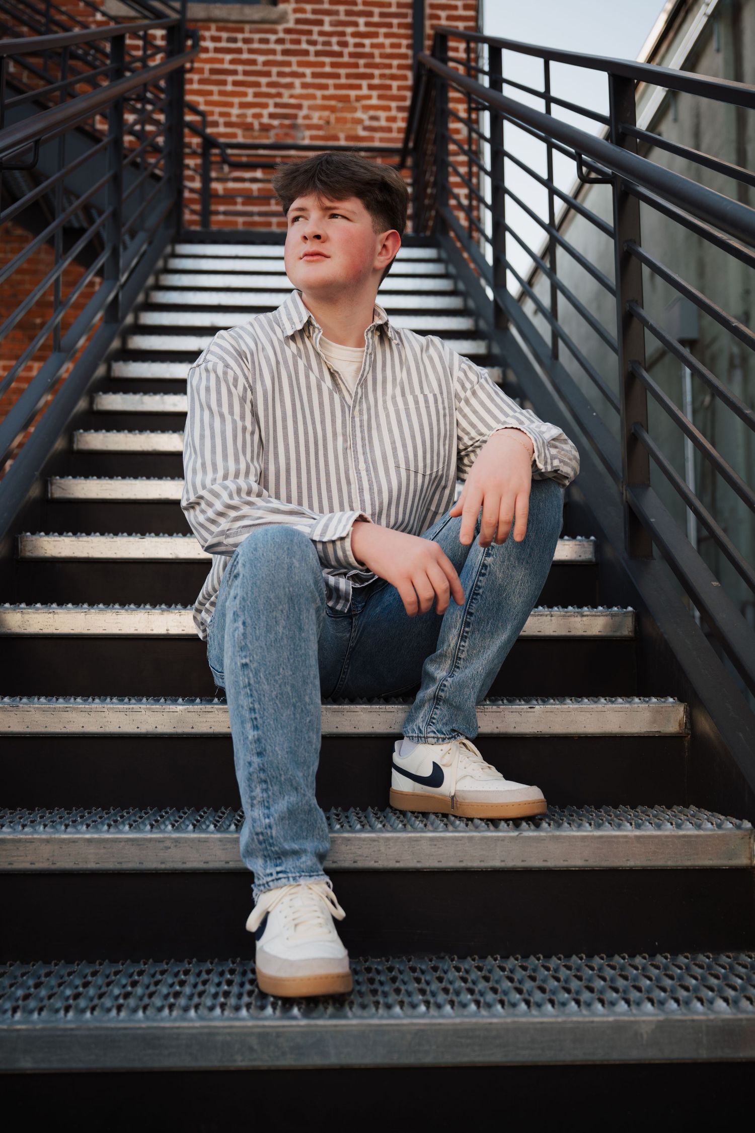 Relaxed portrait on metal stairs wearing striped shirt and jeans with white sneakers.