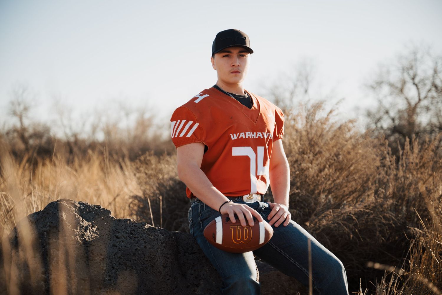 Football player in orange Warriors jersey posing with ball in field setting.
