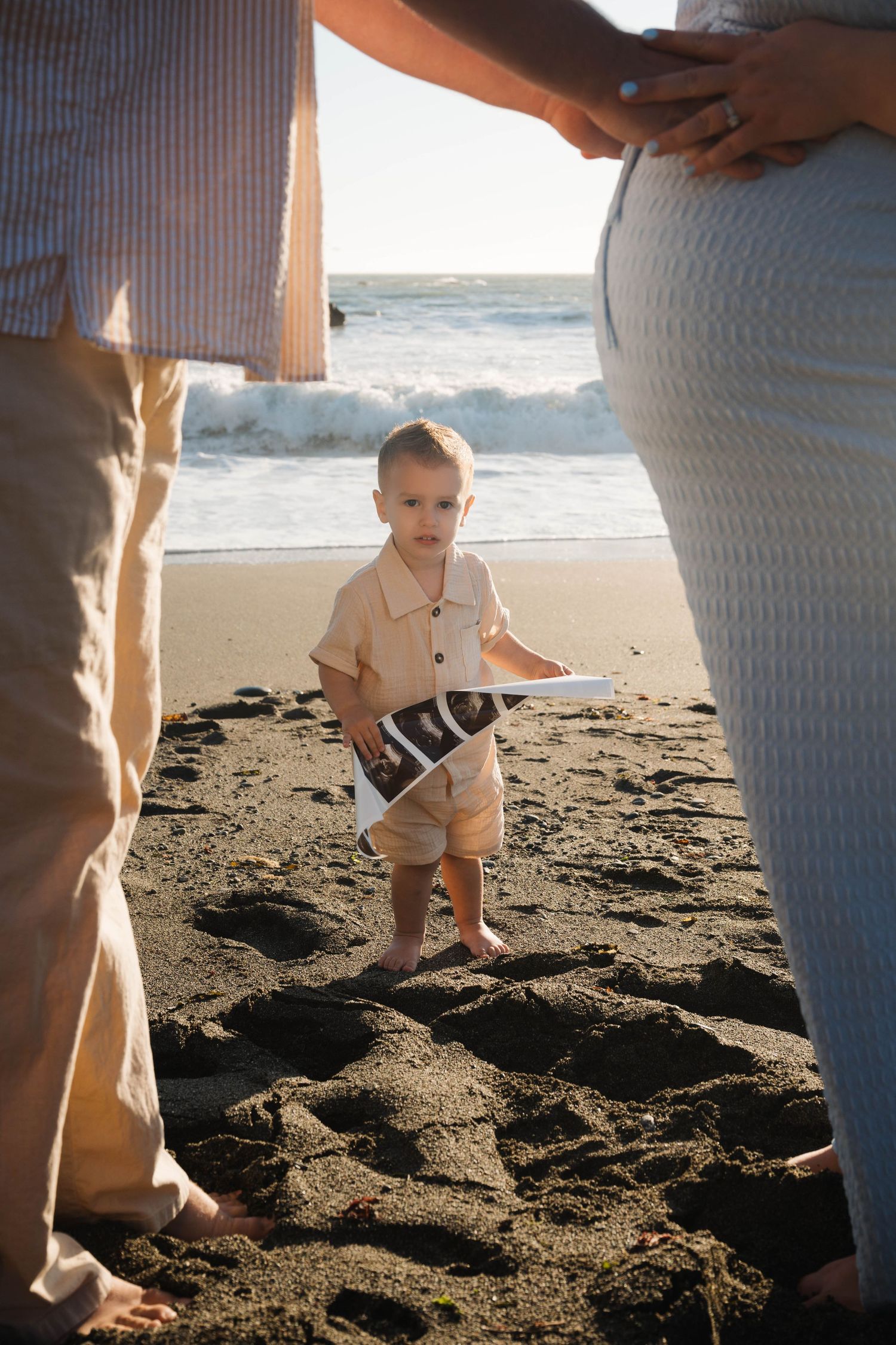 A toddler holds an ultrasound photo on a black sand beach, standing between parents, one visibly pregnant, with ocean waves behind.