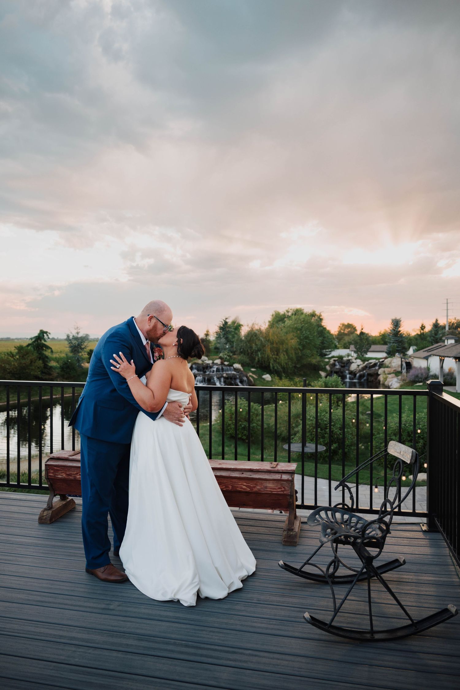 Bride and groom share a romantic kiss on a deck at sunset, with a scenic pond and lush greenery in the background.