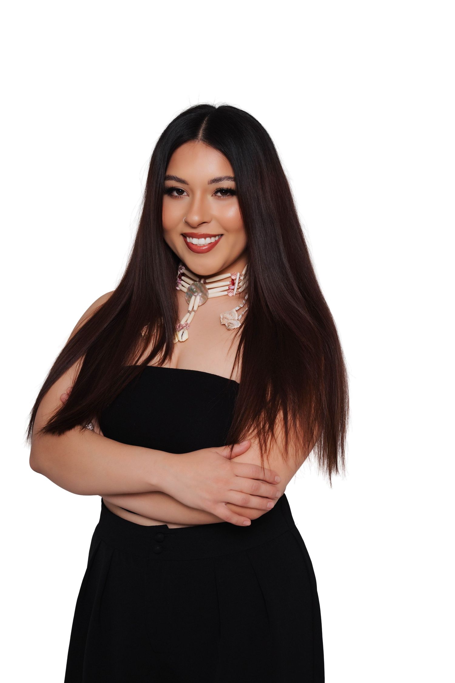 Smiling young woman with long dark hair wearing a black strapless top and floral choker necklace, posing against white background.
