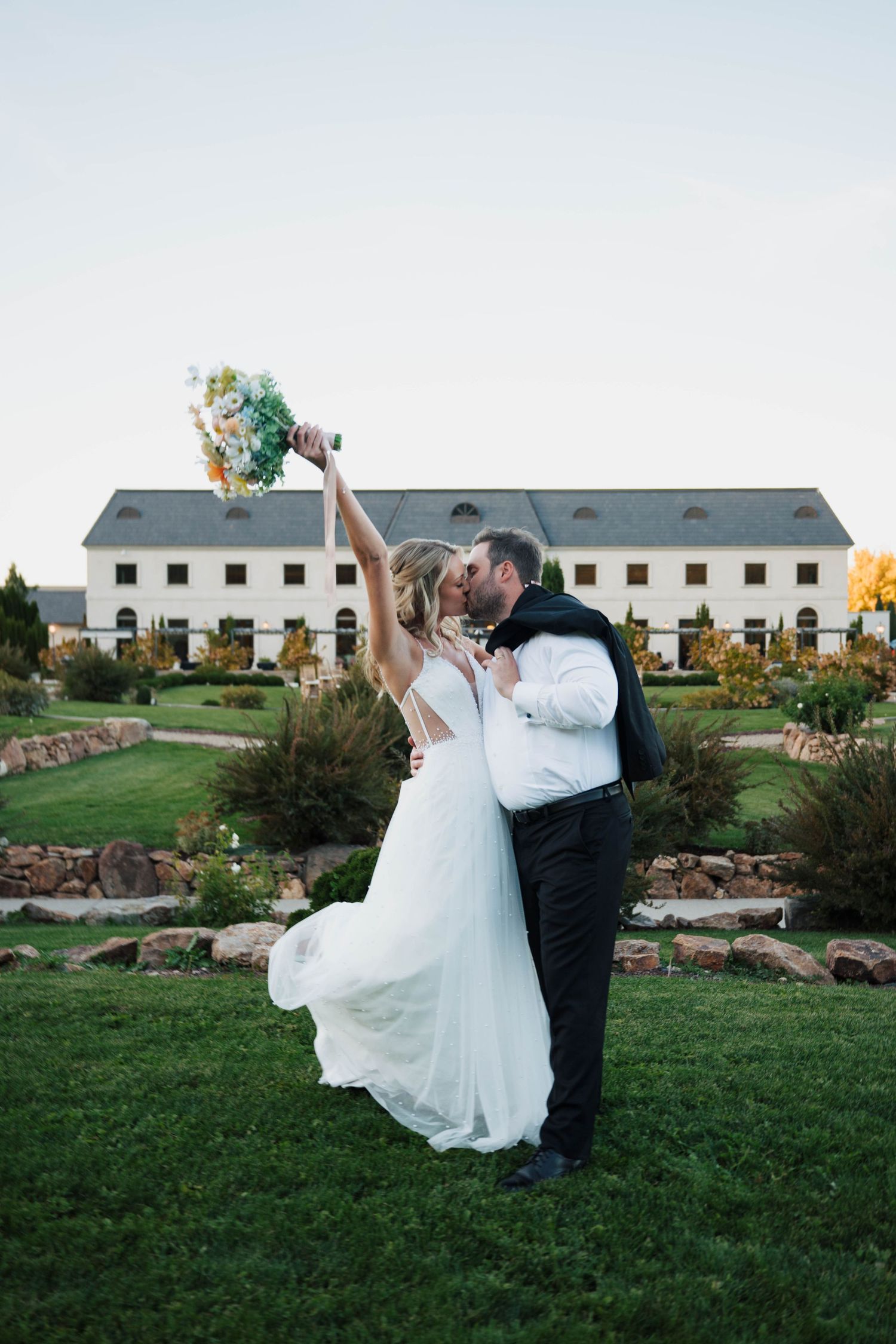 Bride and groom share a romantic kiss on lush green lawn, with bride raising colorful bouquet, elegant white venue behind them.