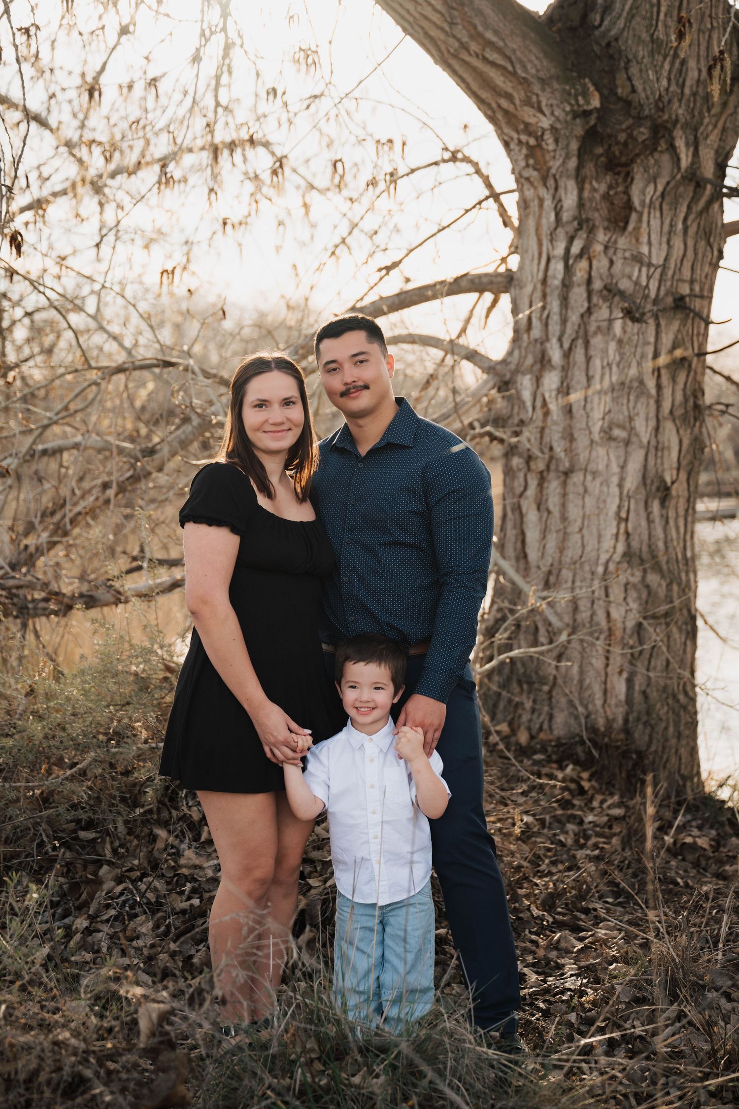 Family of three posing outdoors near a large tree, parents smiling with young son in white shirt at golden hour.