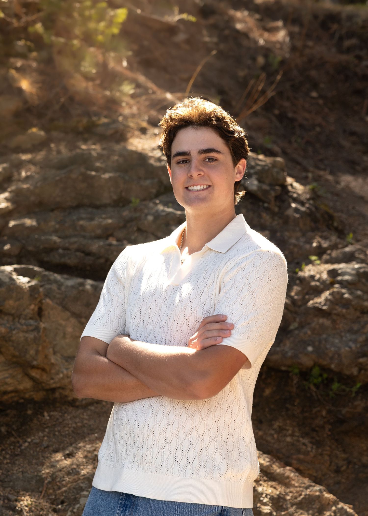 Young man in white knit polo shirt stands arms crossed, smiling outdoors against rocky backdrop in warm sunlight.