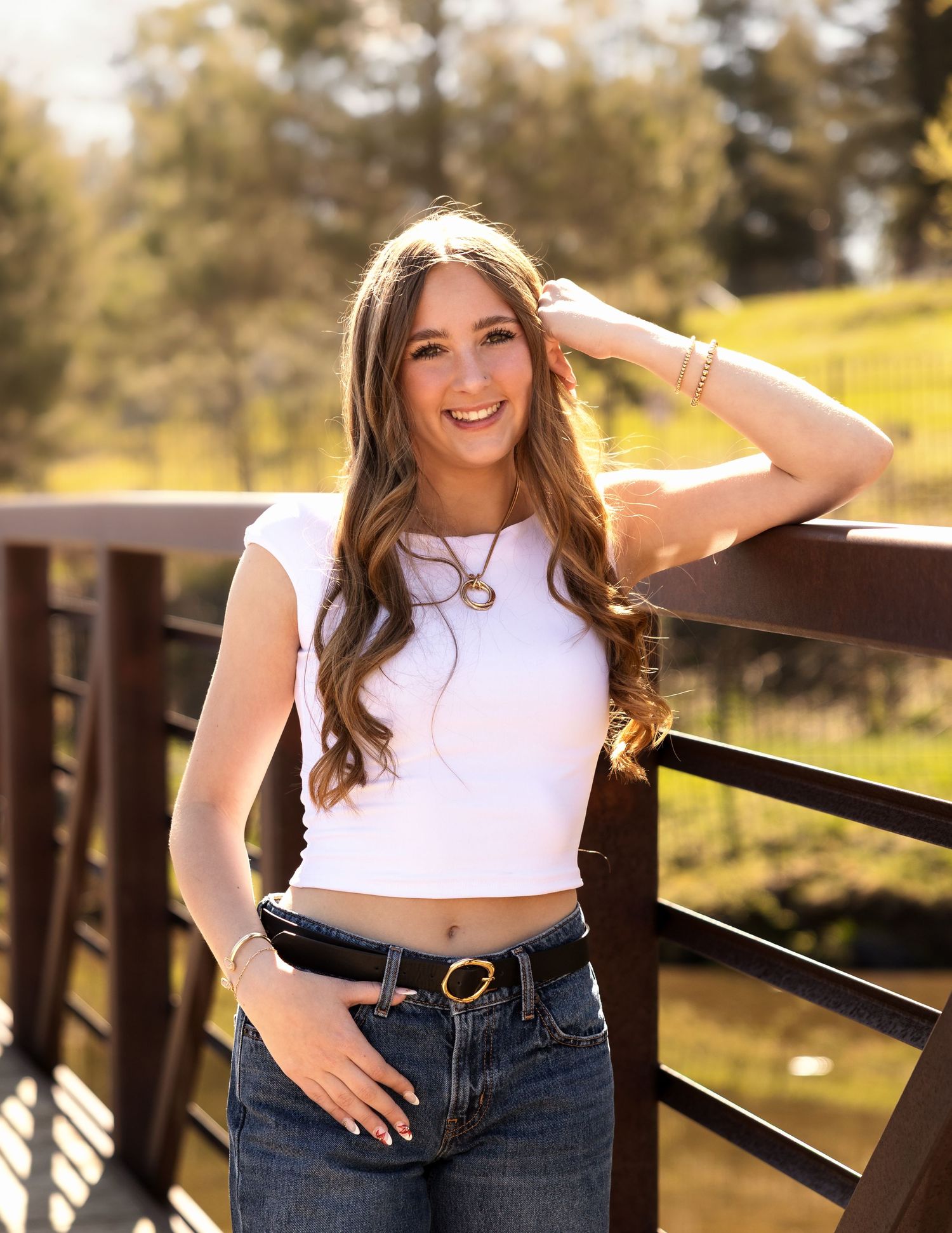 Young woman in white crop top and jeans smiling on a wooden bridge surrounded by greenery in golden hour light.