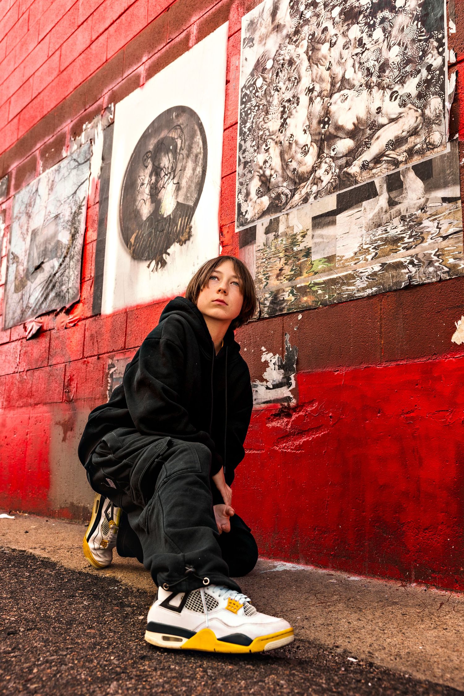 Person crouching in black hoodie and Air Jordan 4 sneakers against a red brick wall with street art.