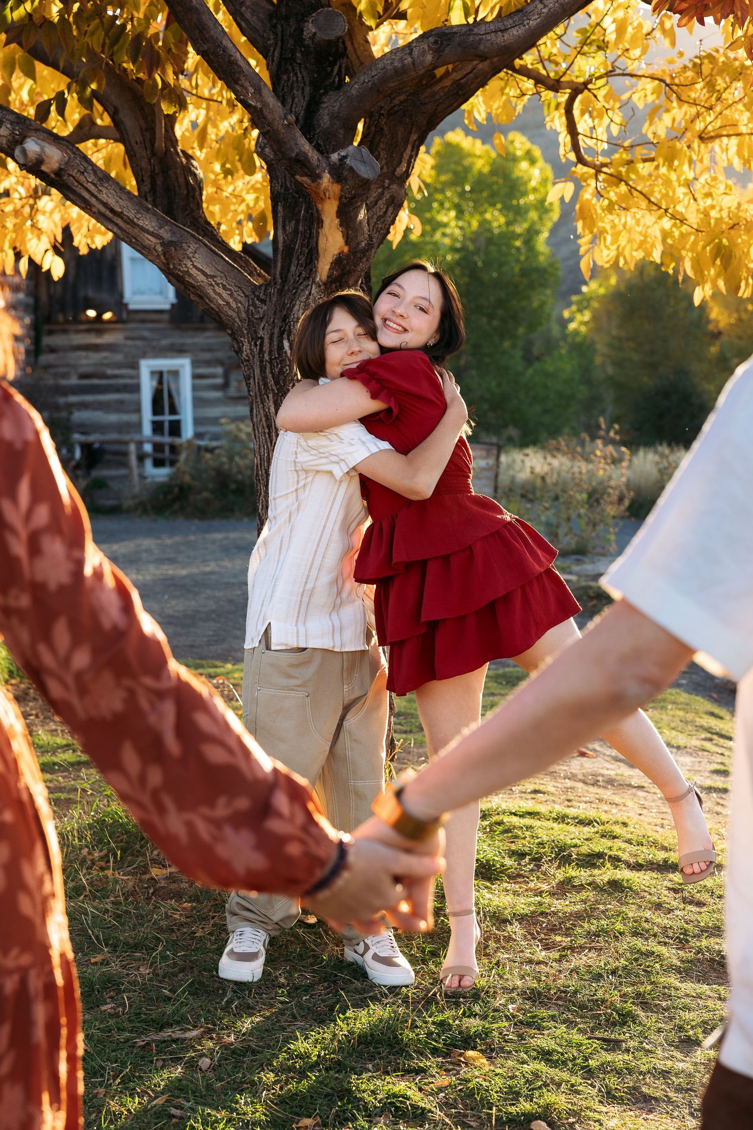 Family photography session with playful teens in an outdoor photoshoot with red dress twirling against golden autumn trees and rustic cabin.