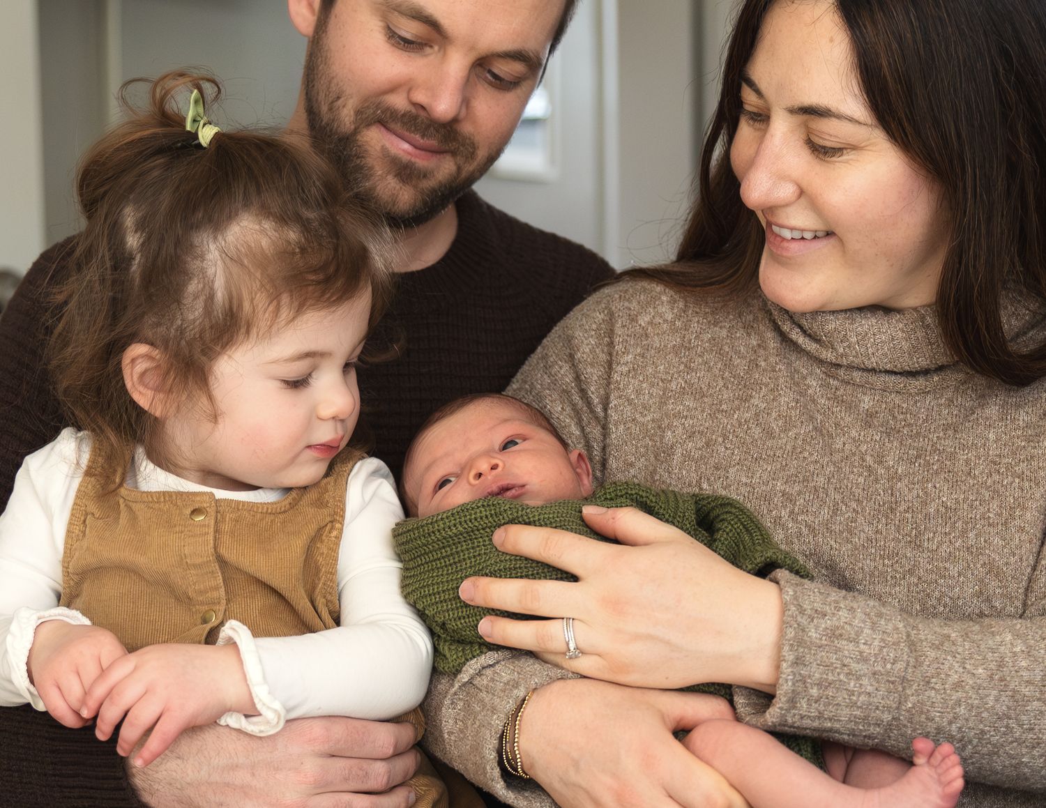 A young family of four with a bearded father, mother, toddler girl, and newborn baby wrapped in green.