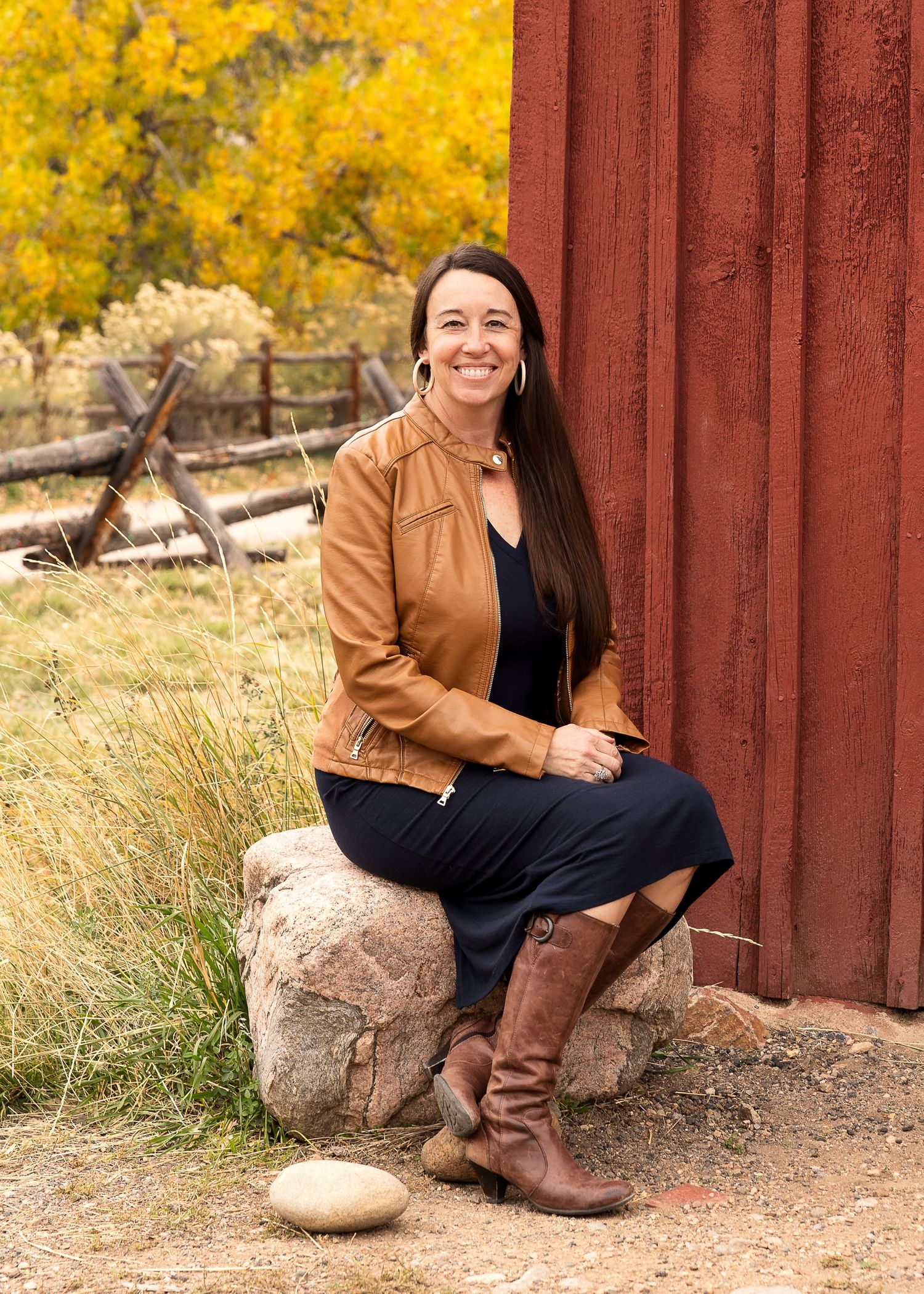 Full body portrait of a woman with a friendly smile sitting on rocks by a red barn with golden foliage in background located in Golden, Co.