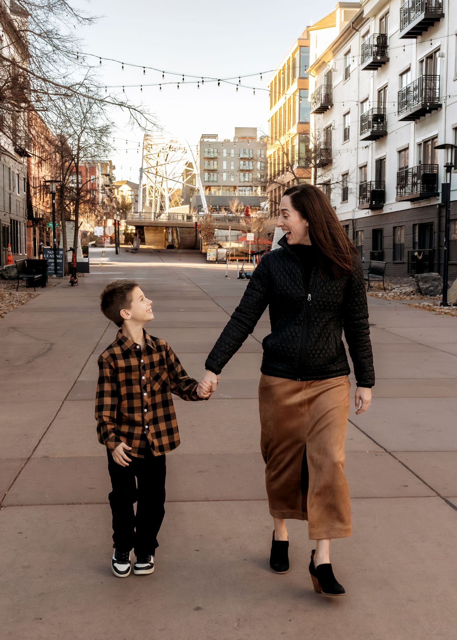 Candid lifestyle photography of a mother and son walking together on an urban street with historic buildings and string lights in Denver, CO