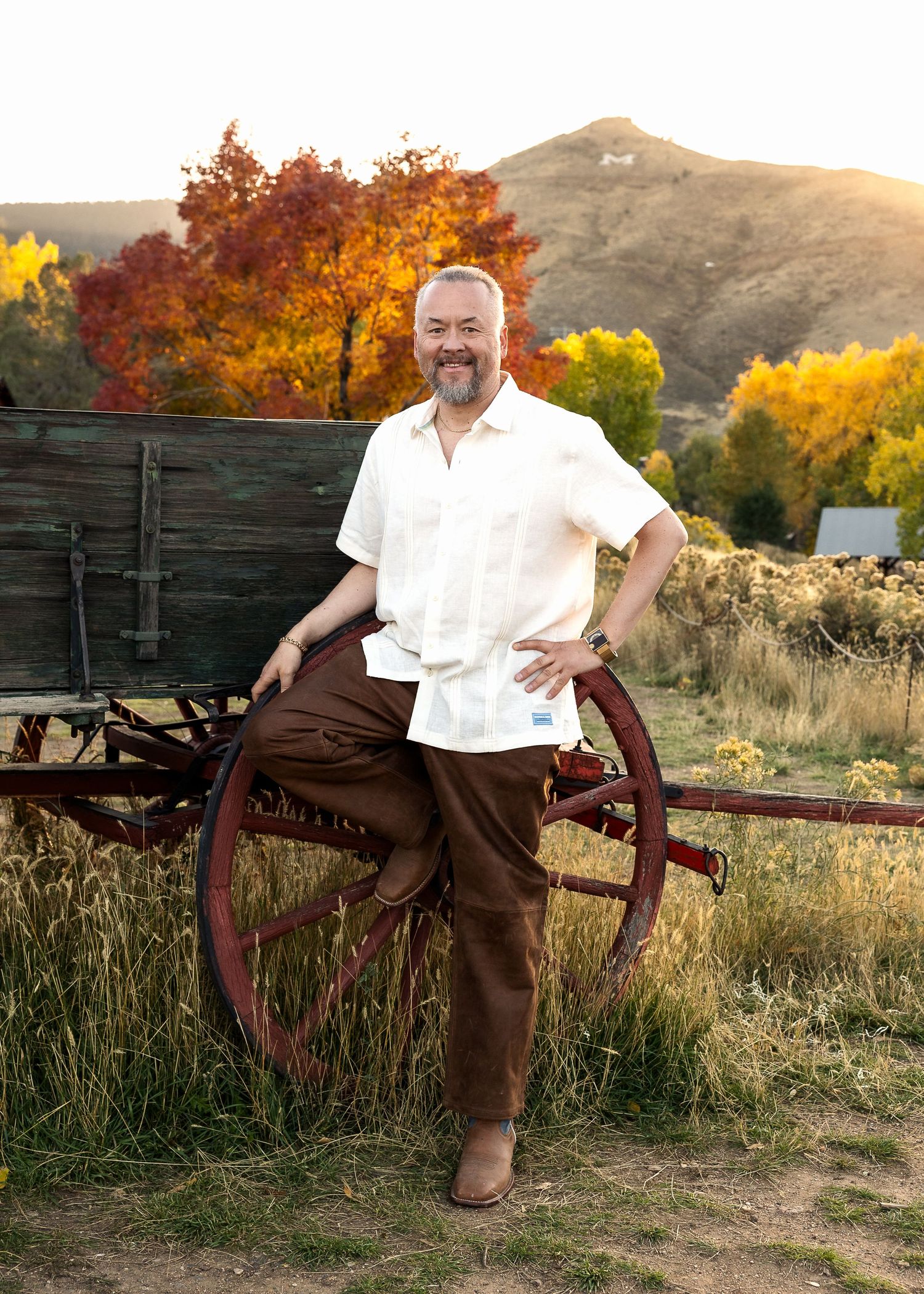 Portrait of man standing by a vintage wooden wagon wheel with autumn foliage and Colorado mountains in background by Arvada, photographer.