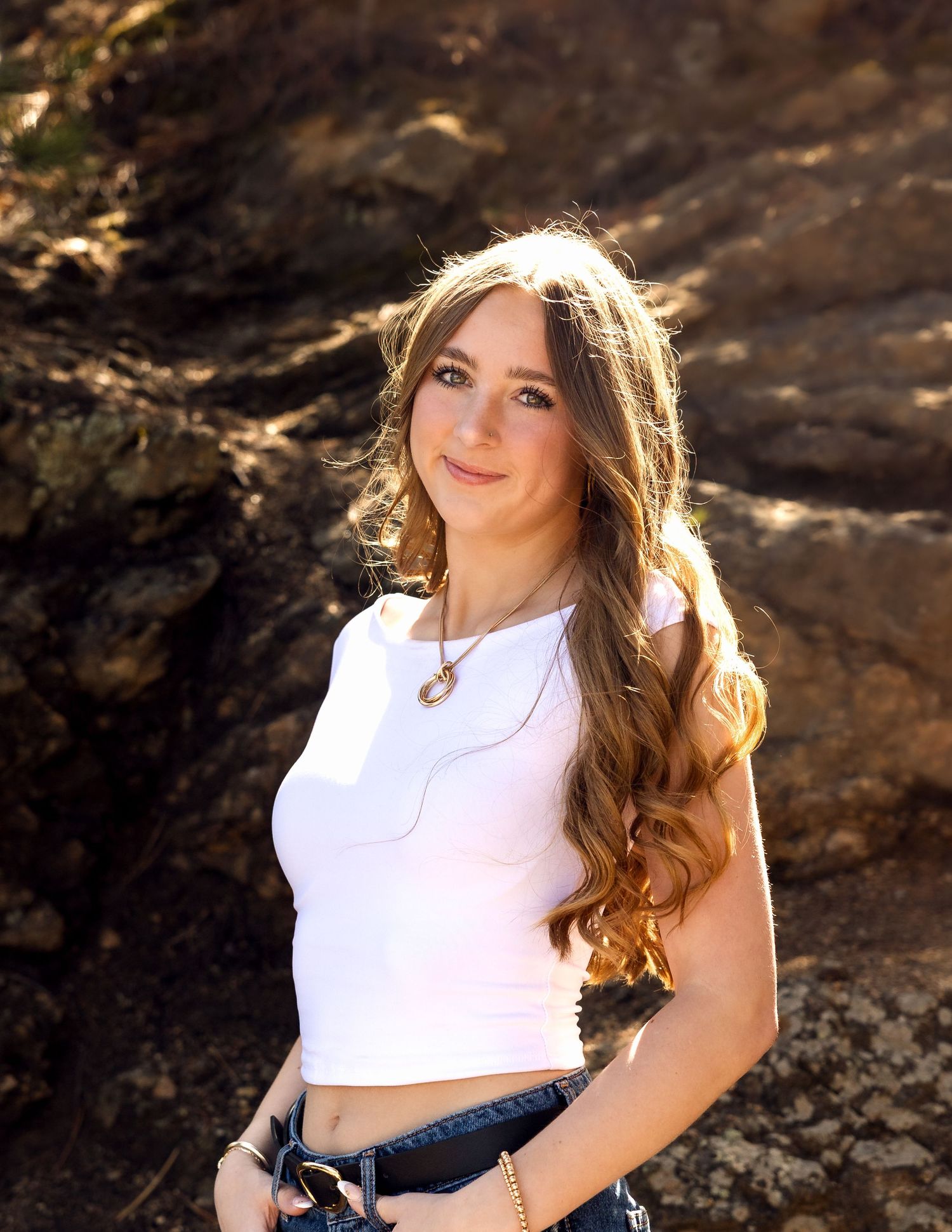Young woman with long wavy hair wearing a white crop top and jeans, posing against rocky background outdoors.