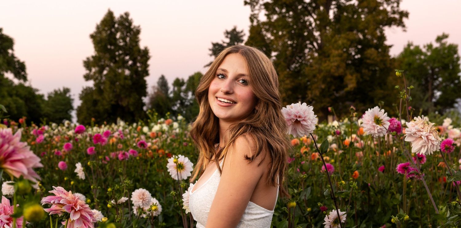 Young woman smiling in a vibrant flower field with pink and white dahlias at sunset.