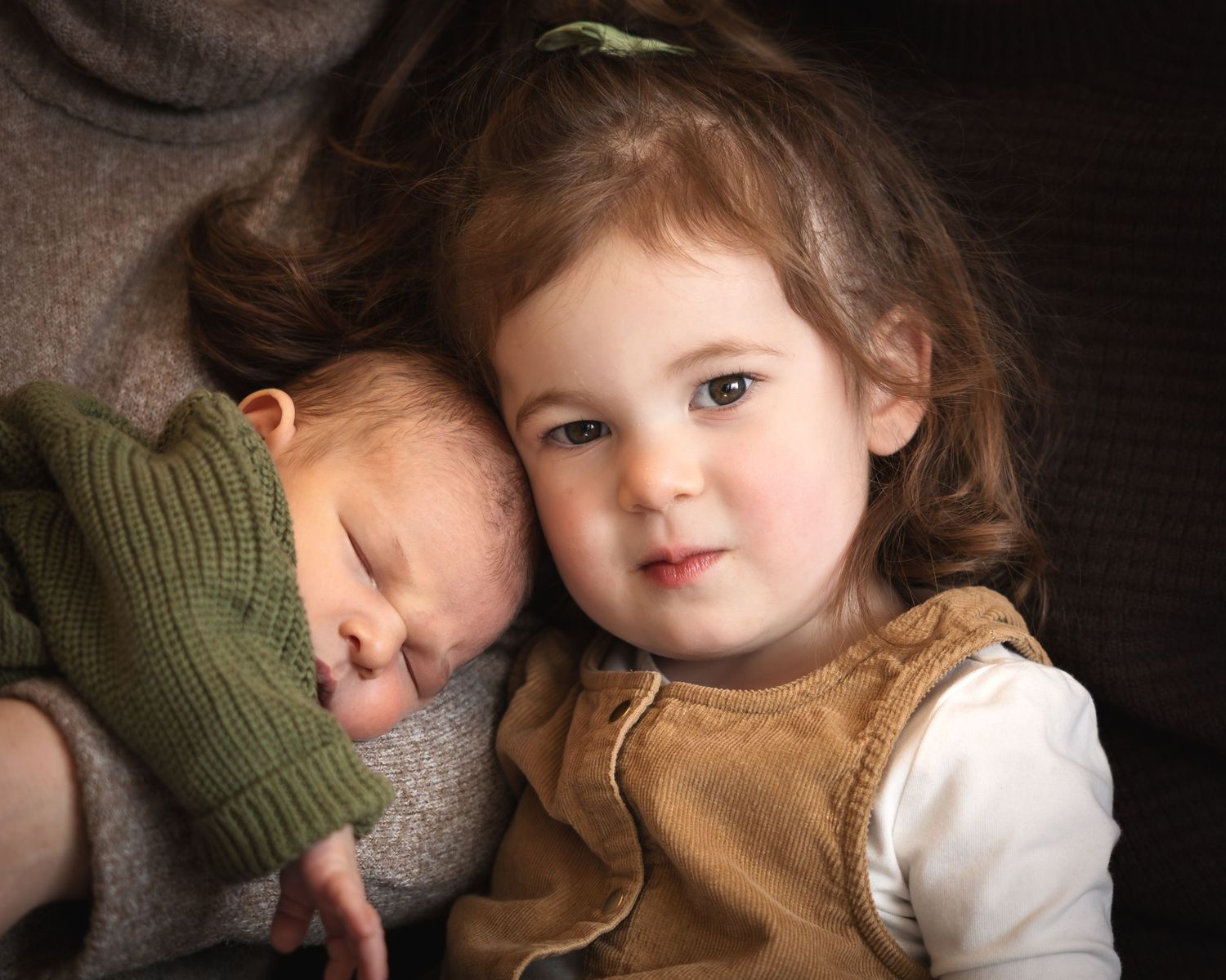 Young girl with curly hair gently holds sleeping newborn baby sibling, wearing green knit hat, in a tender portrait.