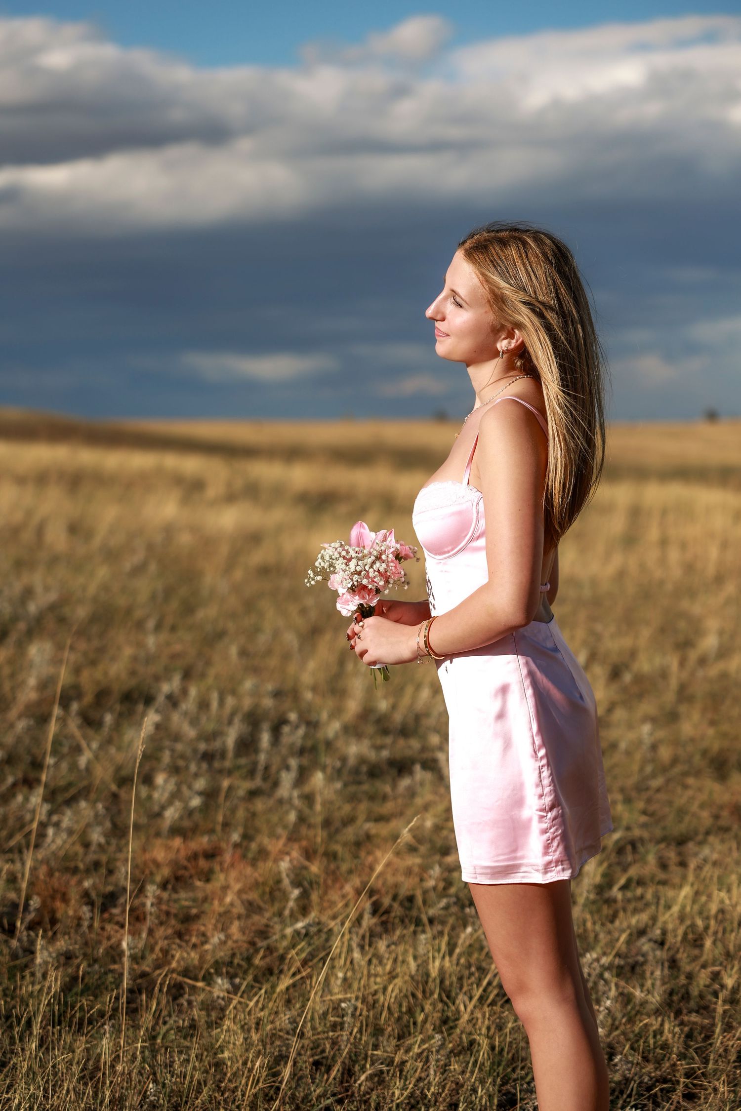 Candid portrait by Wild Light Studio of homecoming girl in pink outfit stands in golden field during sunset.