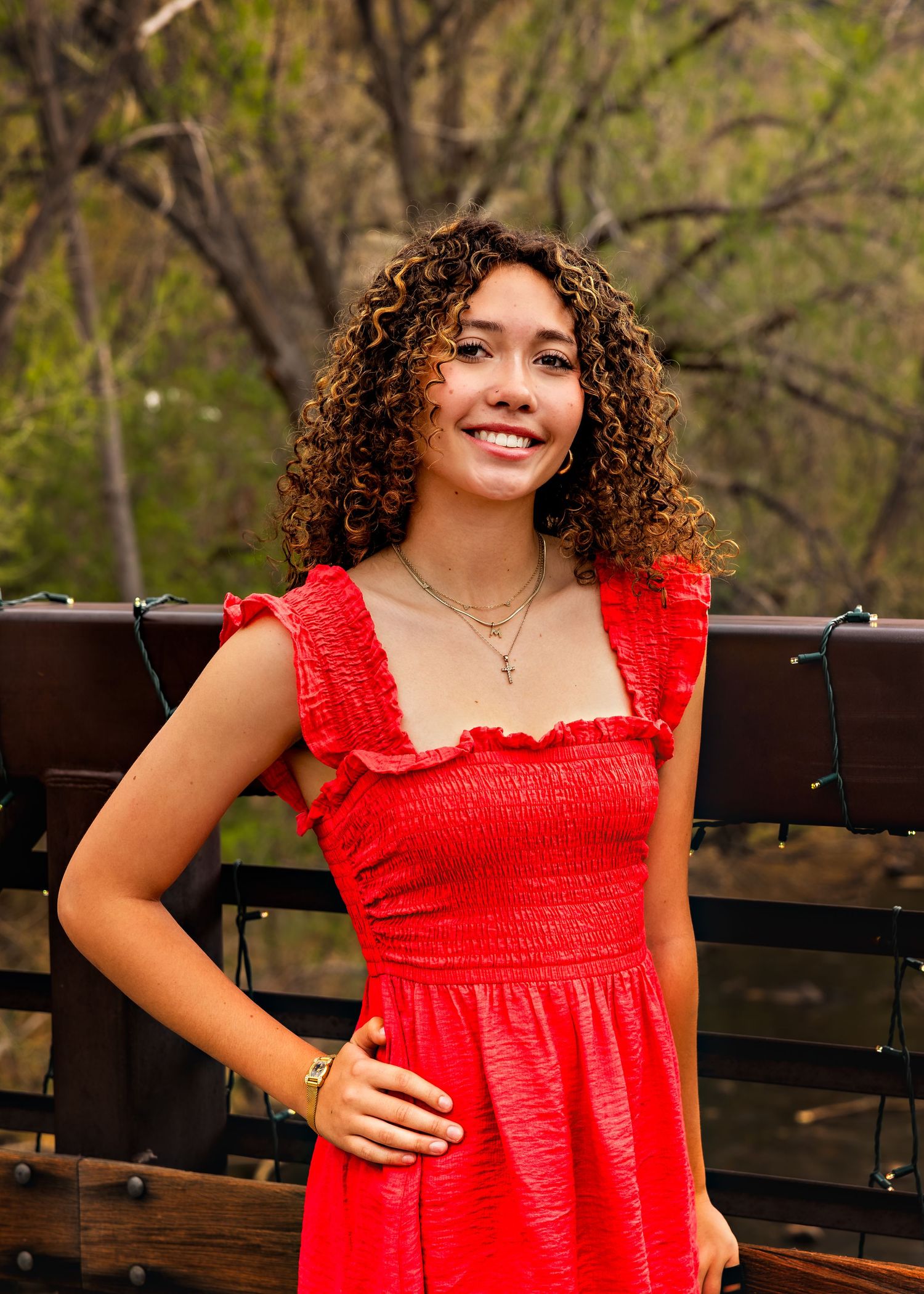Young woman with curly hair smiling in a red ruffled dress, posing outdoors near a wooden fence.