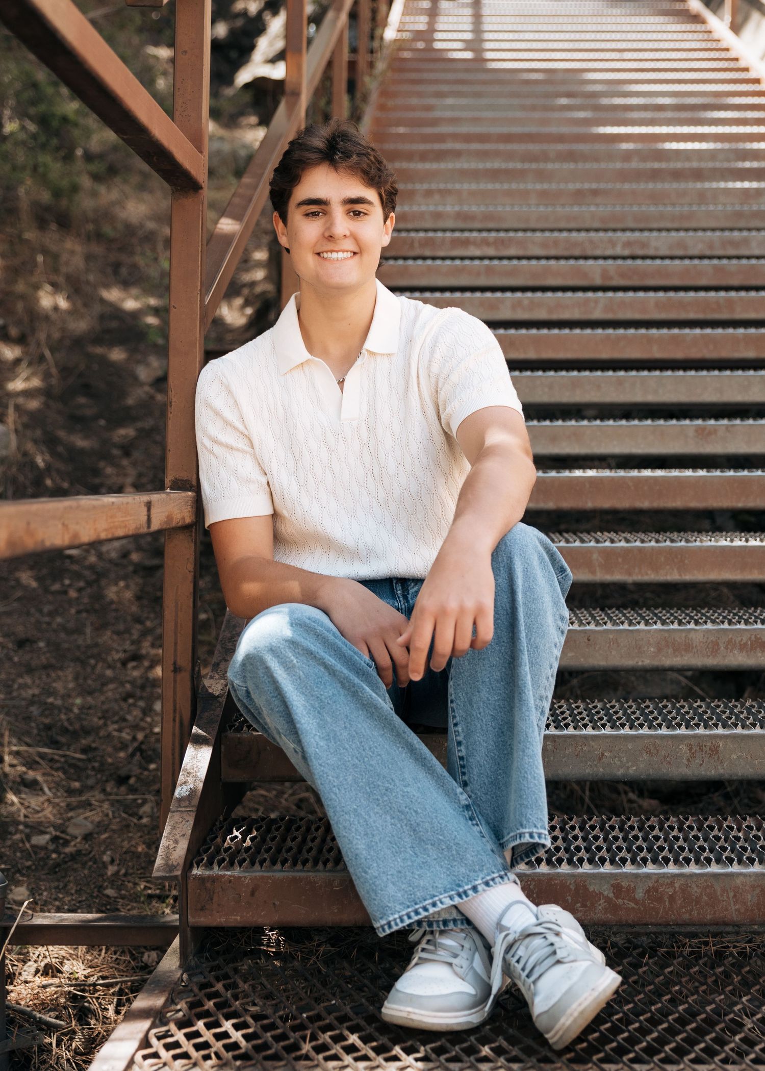Young man in white polo and jeans sitting on rustic metal stairs, smiling outdoors in a senior portrait photo.