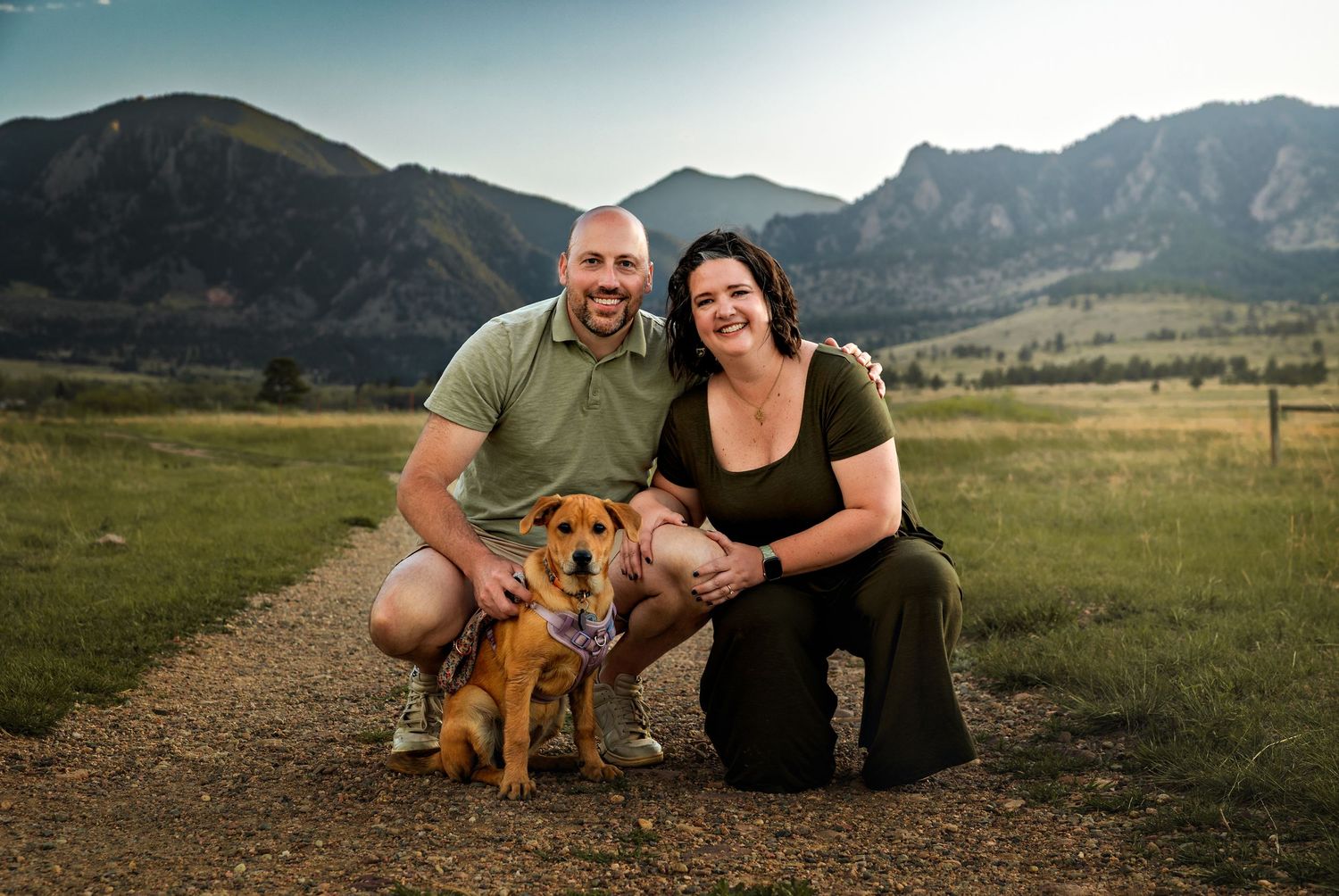 Couple smiling with brown dog in front of scenic Rocky Mountain landscape on sunny day.