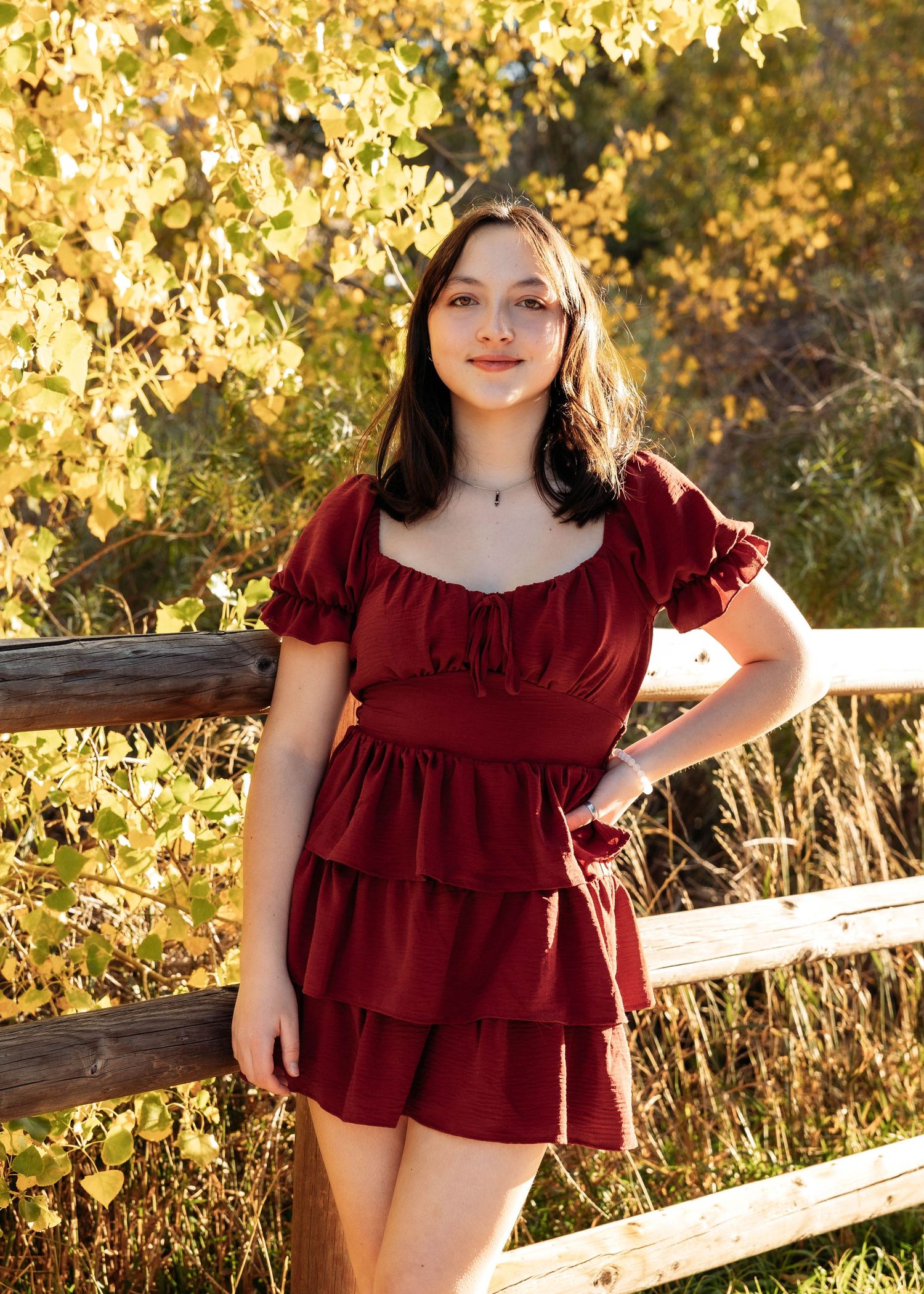 Young woman in a red ruffled mini dress posing by a wooden fence with golden autumn foliage.