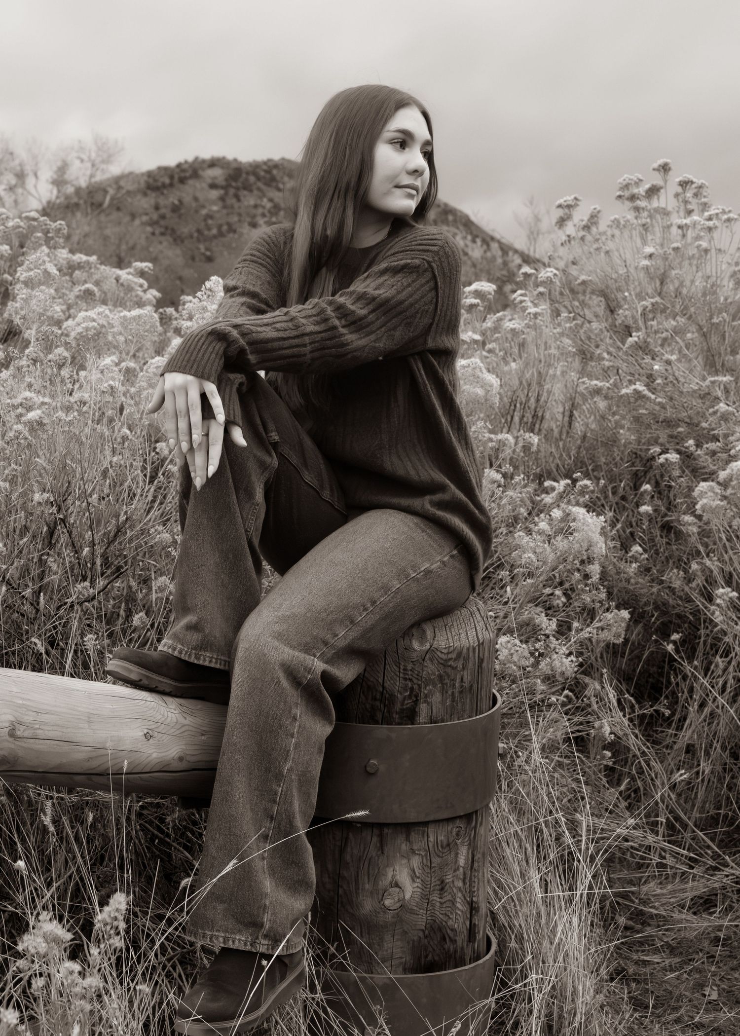 Black and white photo of a young woman sitting on a wooden barrel outdoors, wearing jeans and a sweater.