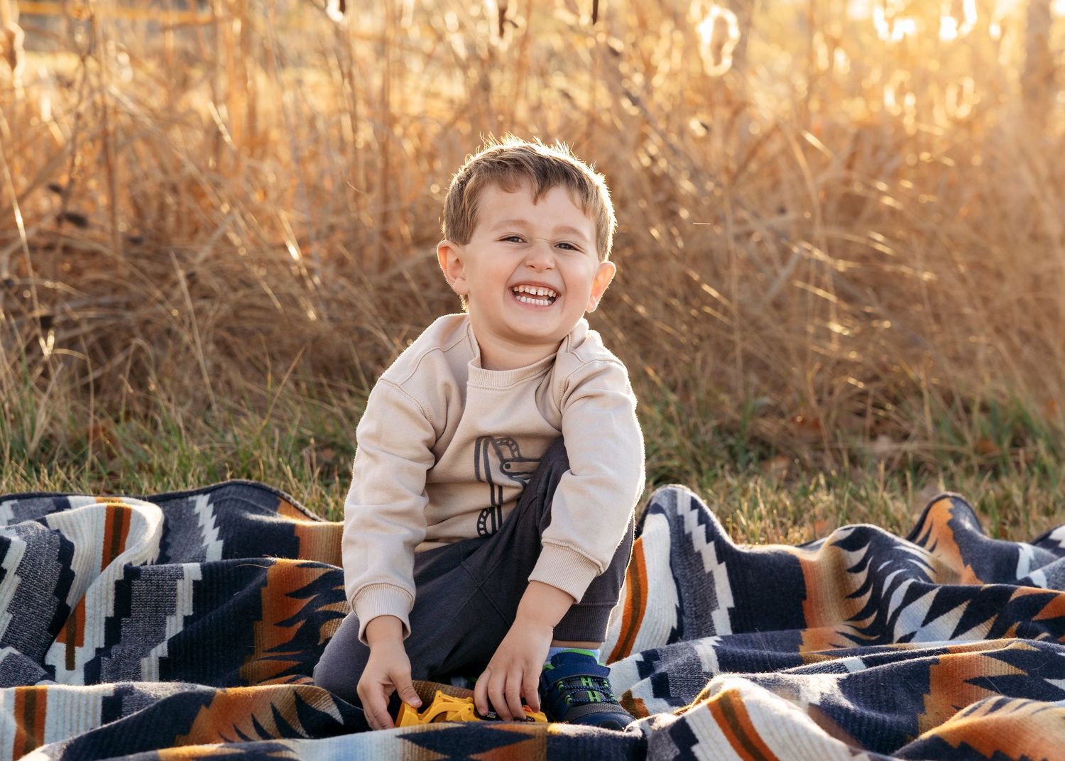 A joyful toddler boy laughs while sitting on a colorful blanket outdoors in a sunny autumn field.