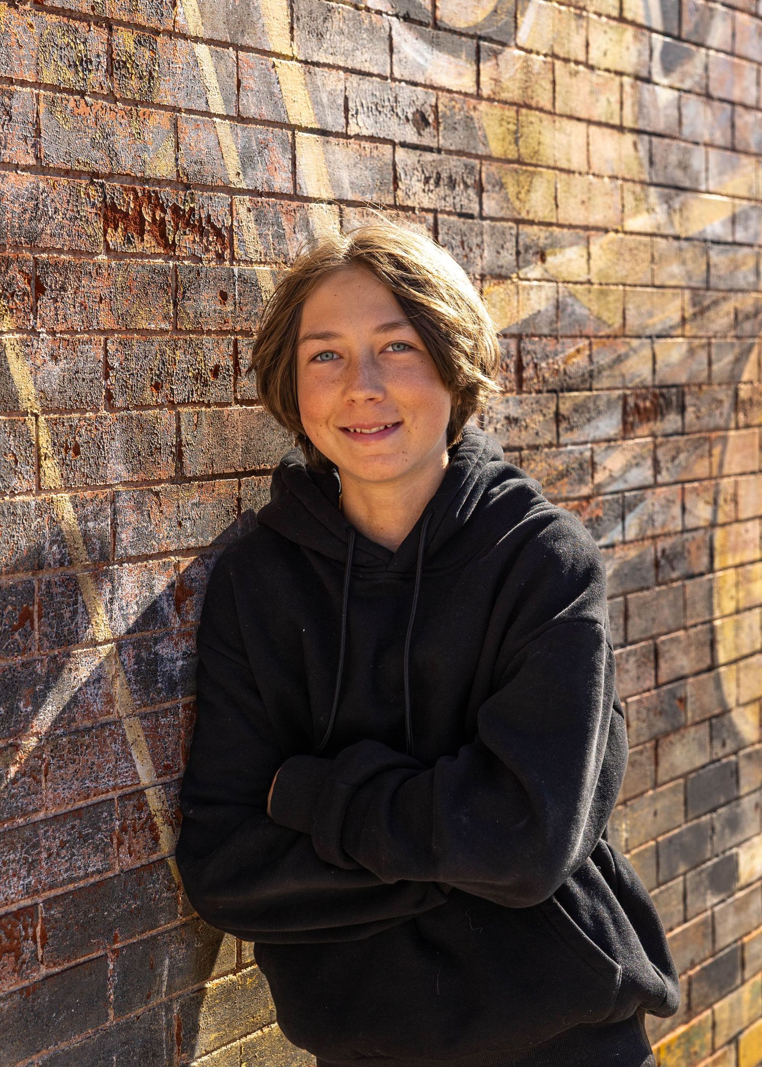 Casual senior portrait of a teen in a black hoodie against a rustic urban brick wall in Denver, CO by Wild Light Studio photographer.