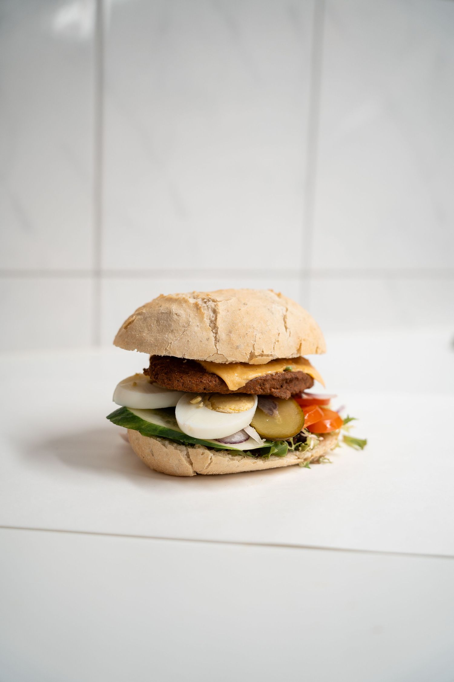 A hearty sandwich on a rustic bun filled with a meat patty, egg, fresh vegetables, and condiments on a white surface.