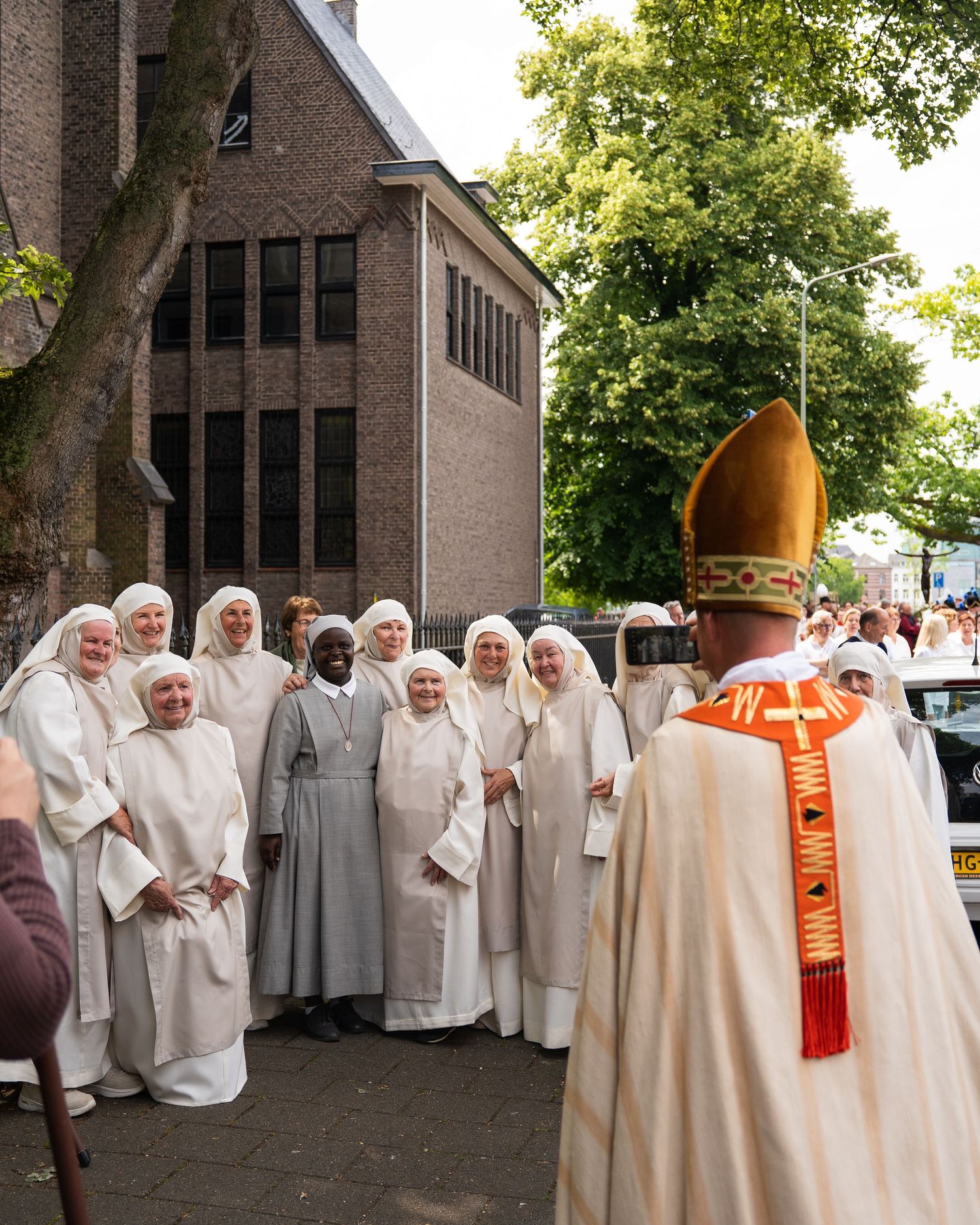 Bishop in orange mitre greets smiling group of nuns in white habits outdoors.