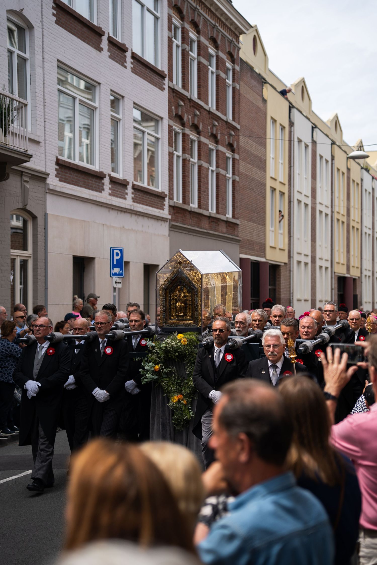 Men in black formal attire with white gloves carry golden reliquary shrine through city streets.