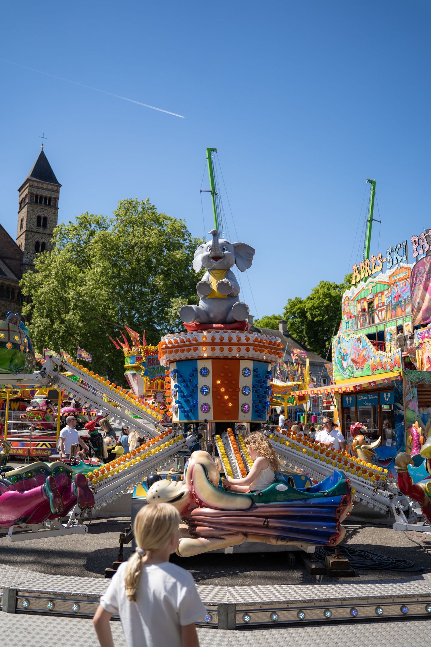Children enjoy a colorful elephant-themed carousel ride at a sunny outdoor fairground with a church in the background.