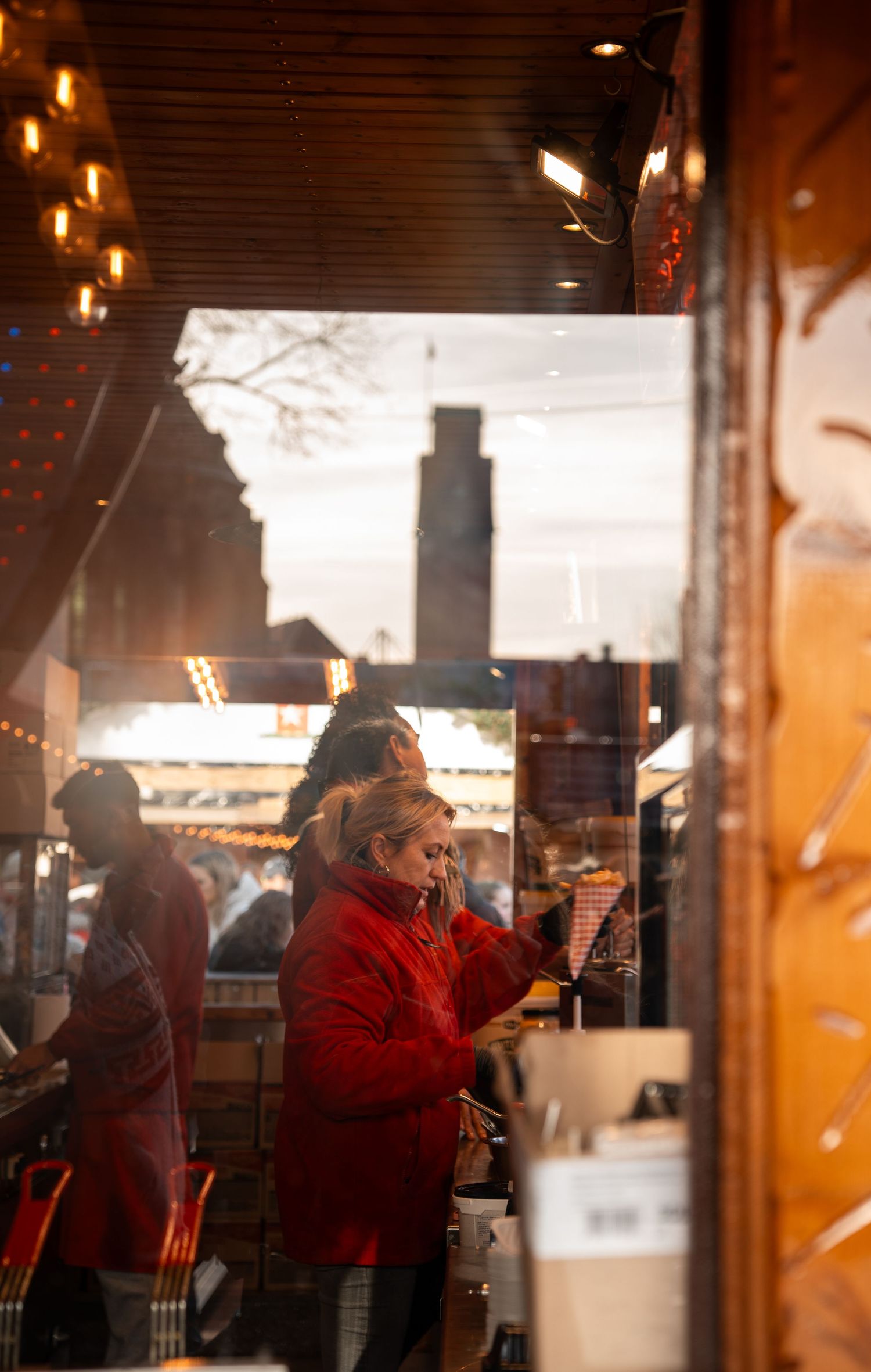 Woman in red coat working at a warmly lit Christmas market stall with festive decorations.