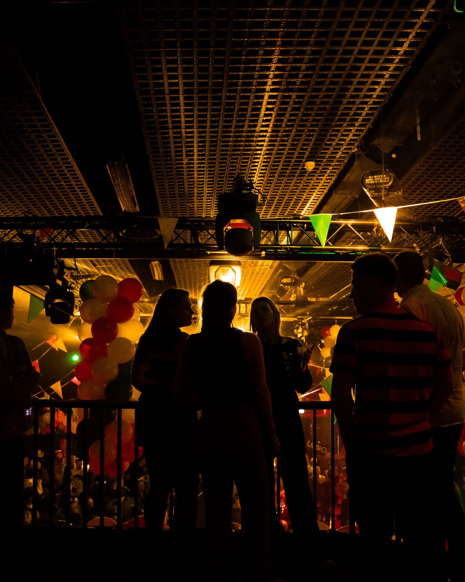 Silhouettes of people watching a party event with balloons and colorful lights in a dark venue.