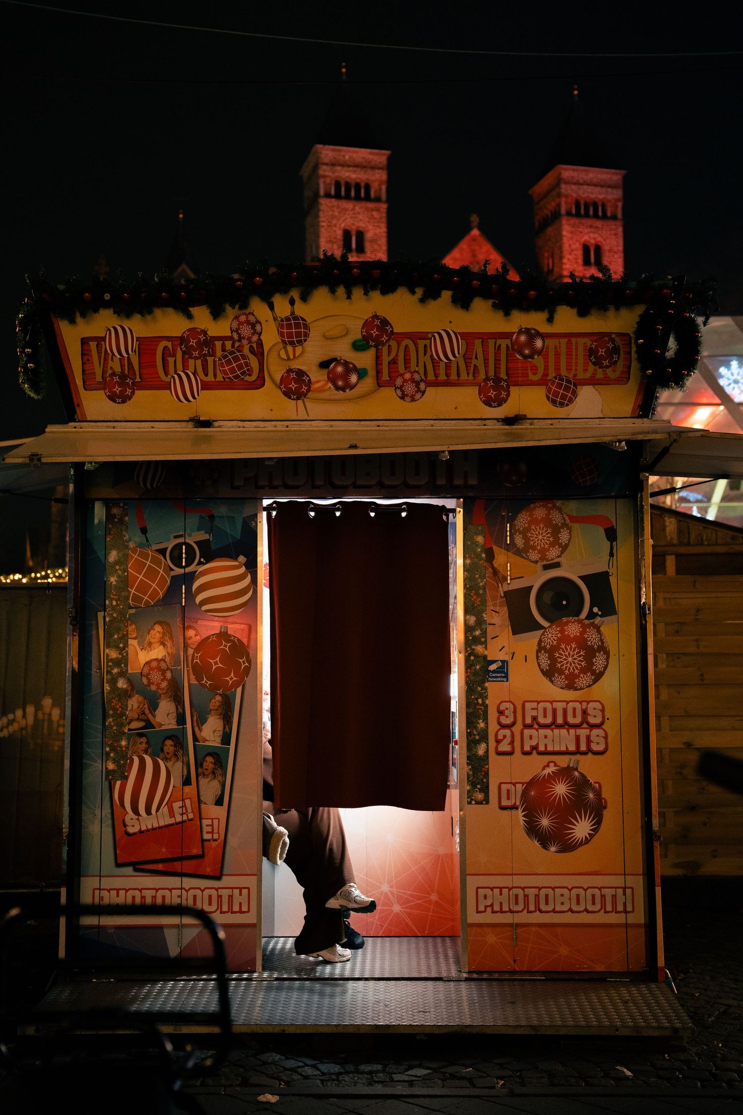 Person inside a colorful vintage photobooth at a night fair with illuminated church towers in background.