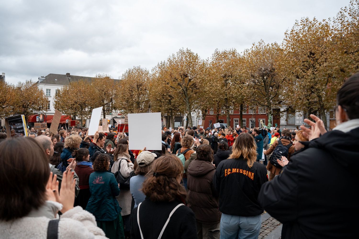 Large crowd gathered at an outdoor protest rally with signs and banners on an overcast autumn day.