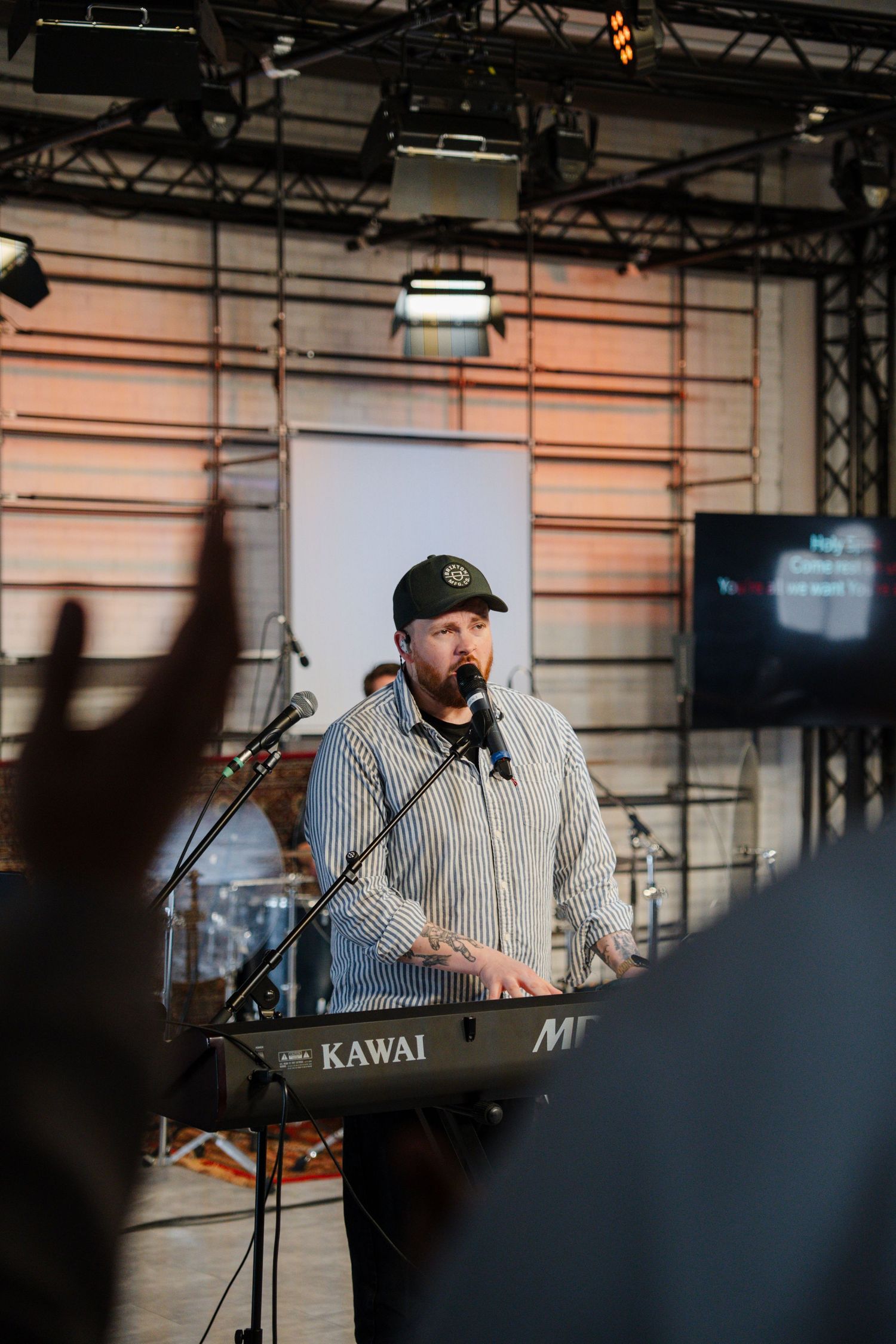 Male worship leader playing a Kawai keyboard and singing into a microphone on a lit church stage.