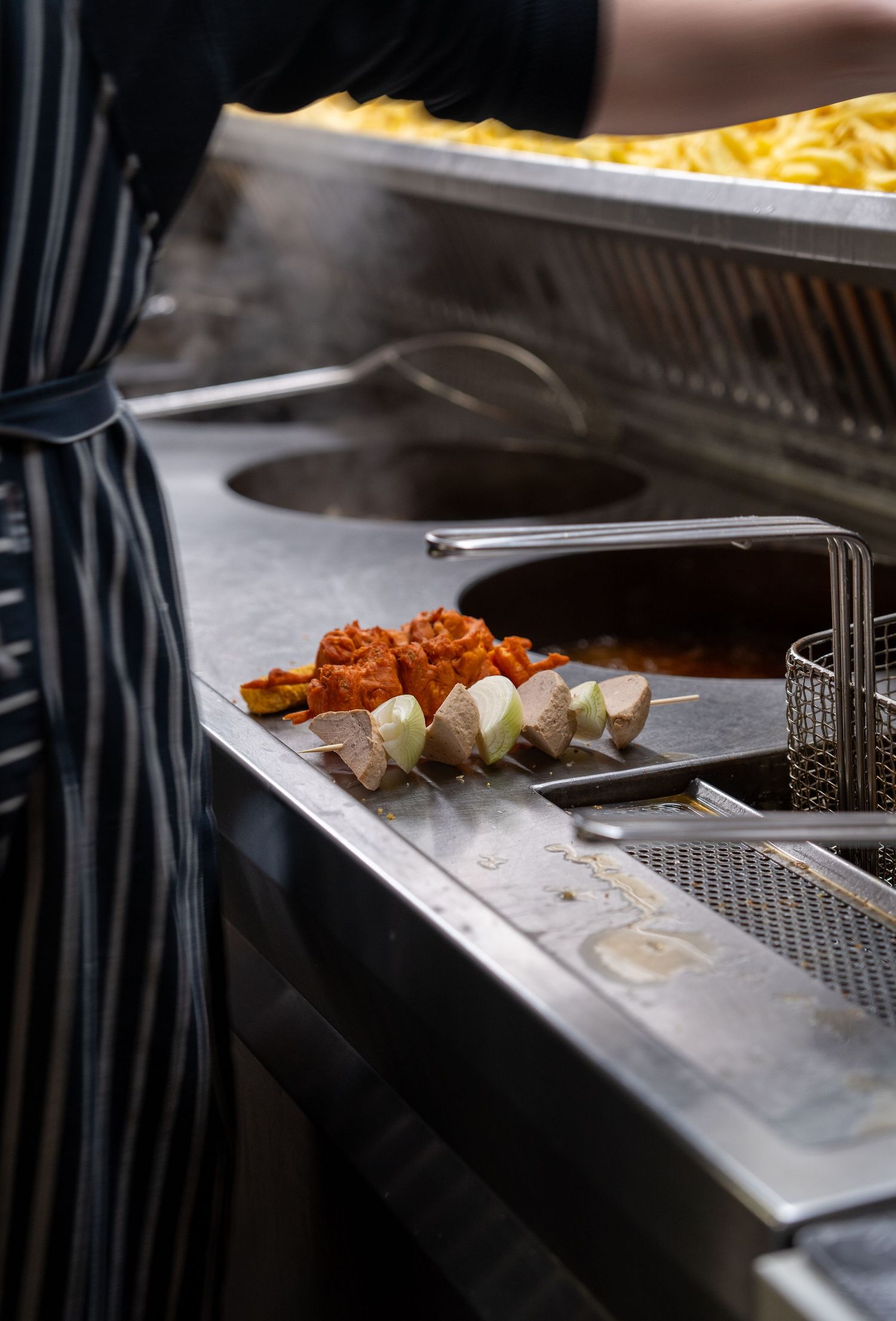 Chef preparing skewers with marinated meat and vegetables on a stainless steel commercial kitchen counter with fries.