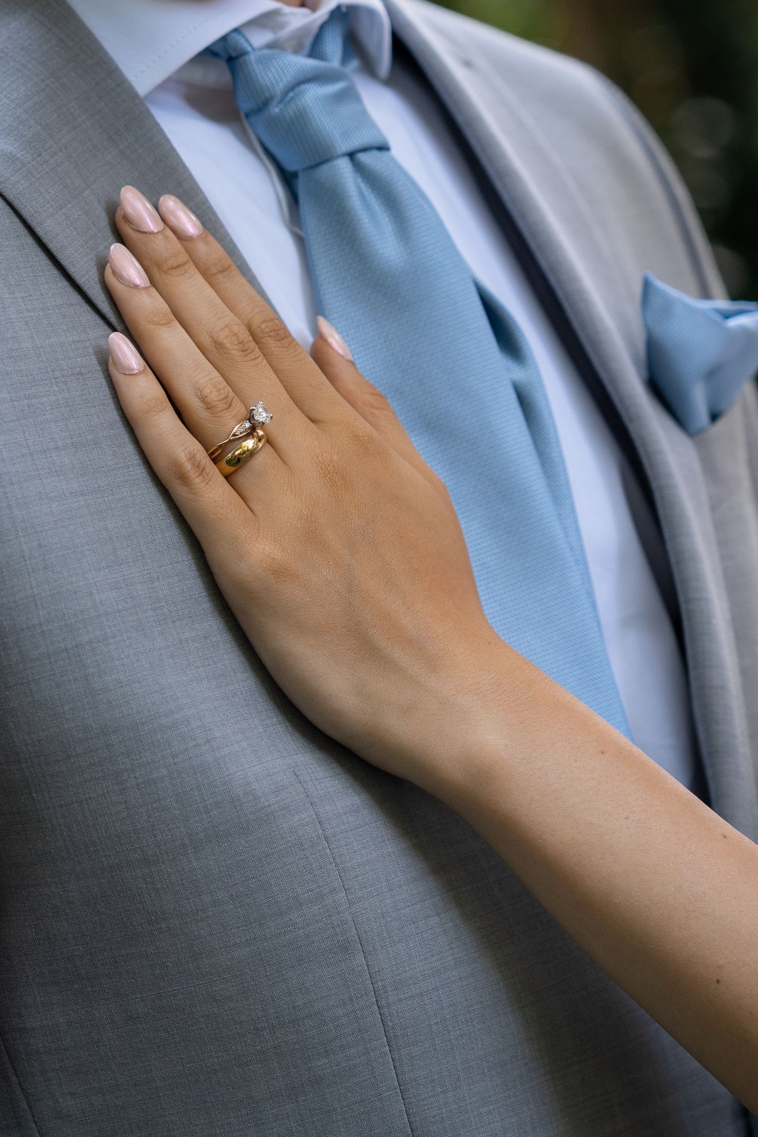 Bride's hand with gold wedding ring resting on groom's gray suit jacket with blue tie.